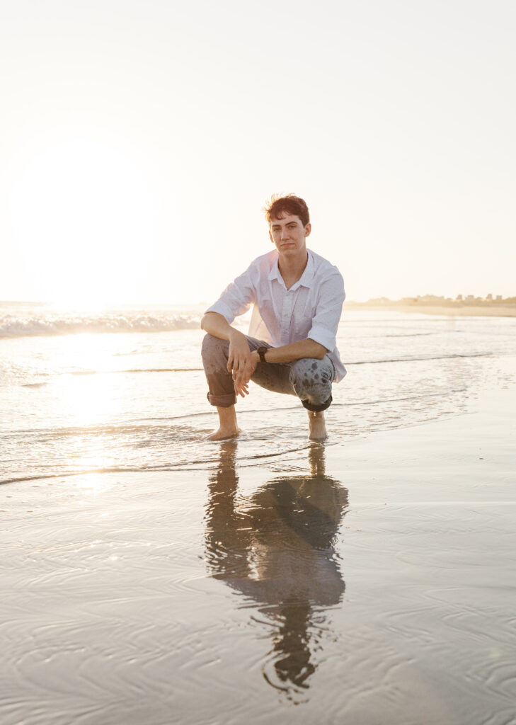 A teenage boy squats in the sand at the beach, looking towards the photographer taking his photo.