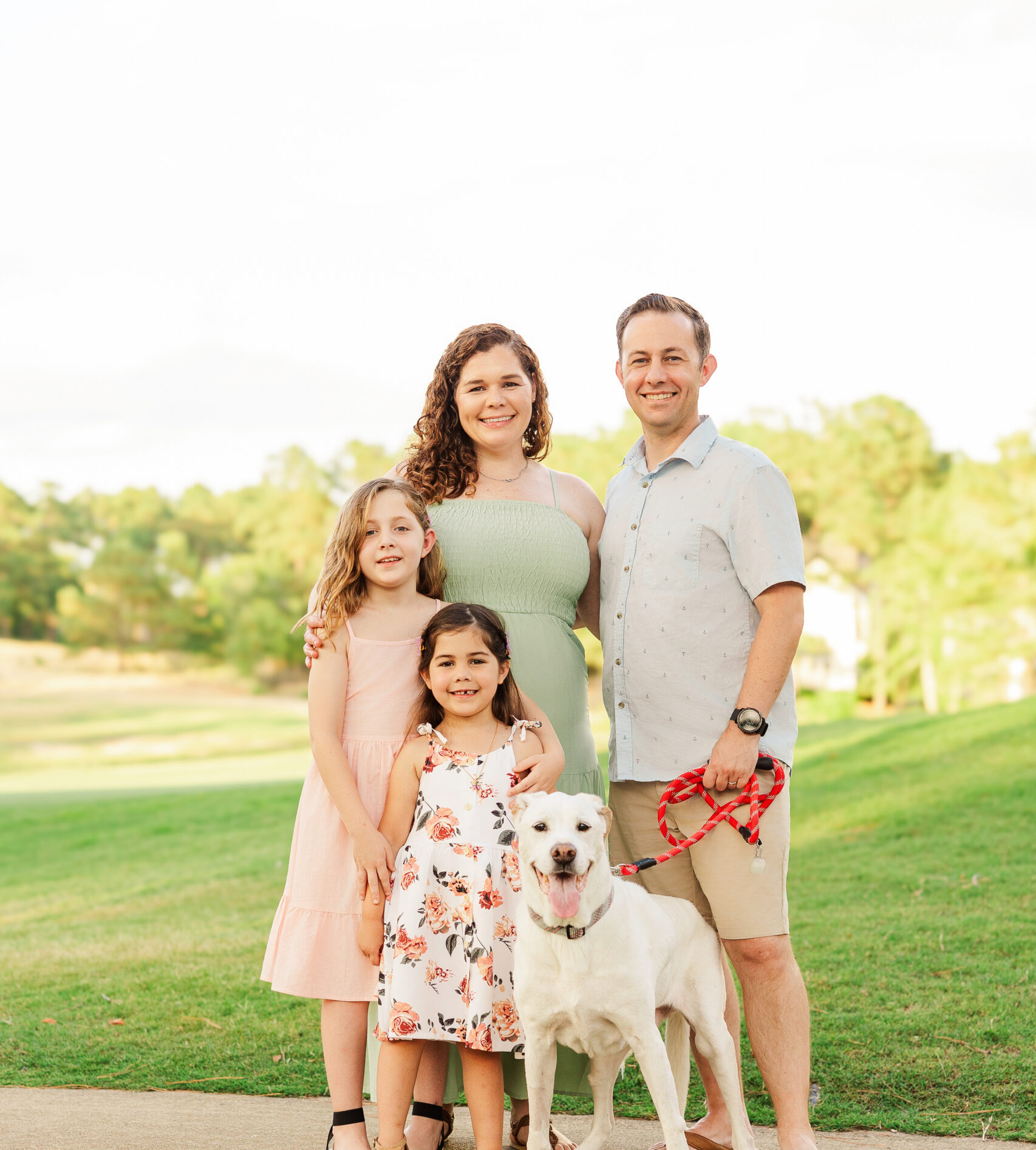 A man and woman stand closely together, smiling with their two young daughters and a white dog on a leash. They are outdoors on a paved pathway with a grassy field and trees in the background. The children are wearing dresses, and the man is in shorts.