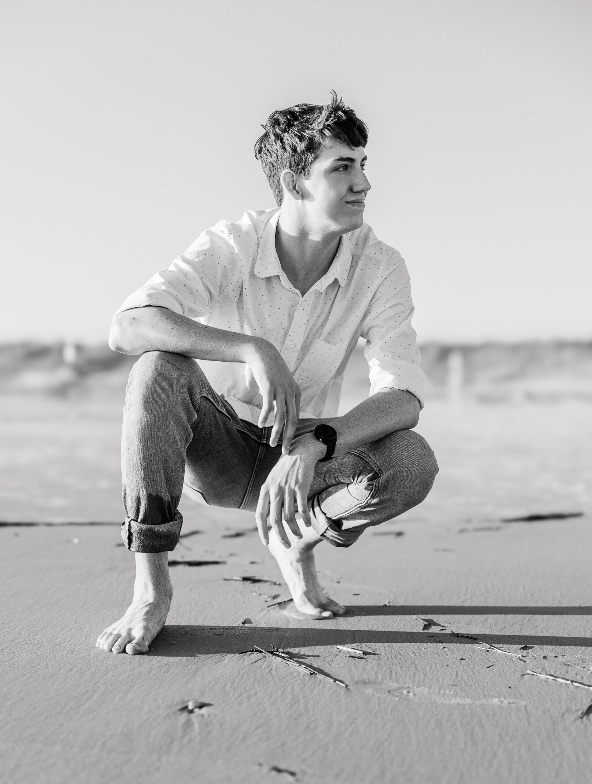 A teenage boy squats in the sand at the beach, looking away from the camera during his high school senior photo session.