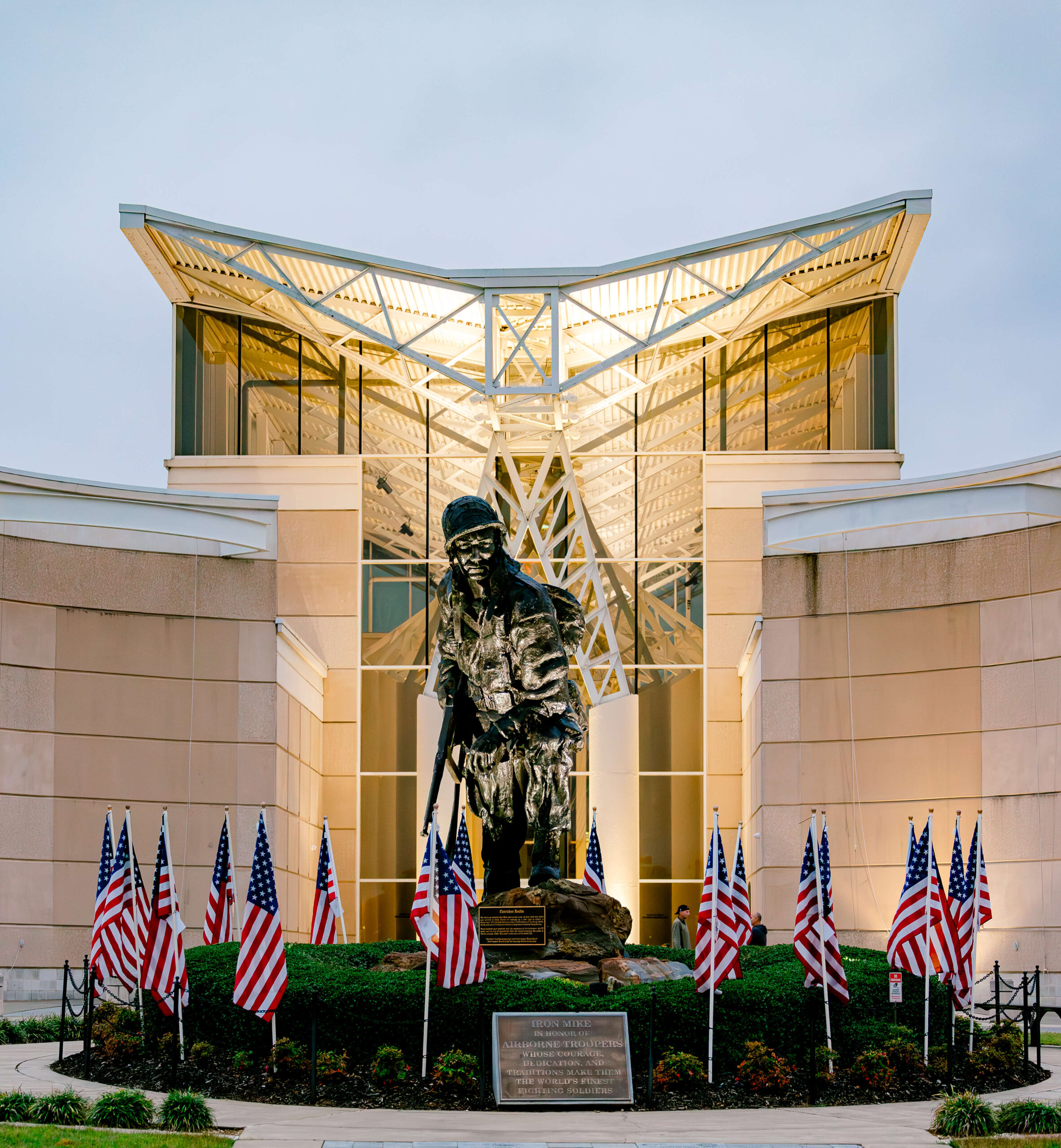 A statue of a soldier stands in front of a modern building with a glass facade. The statue is surrounded by a garden of greenery and several American flags. The sky appears overcast.