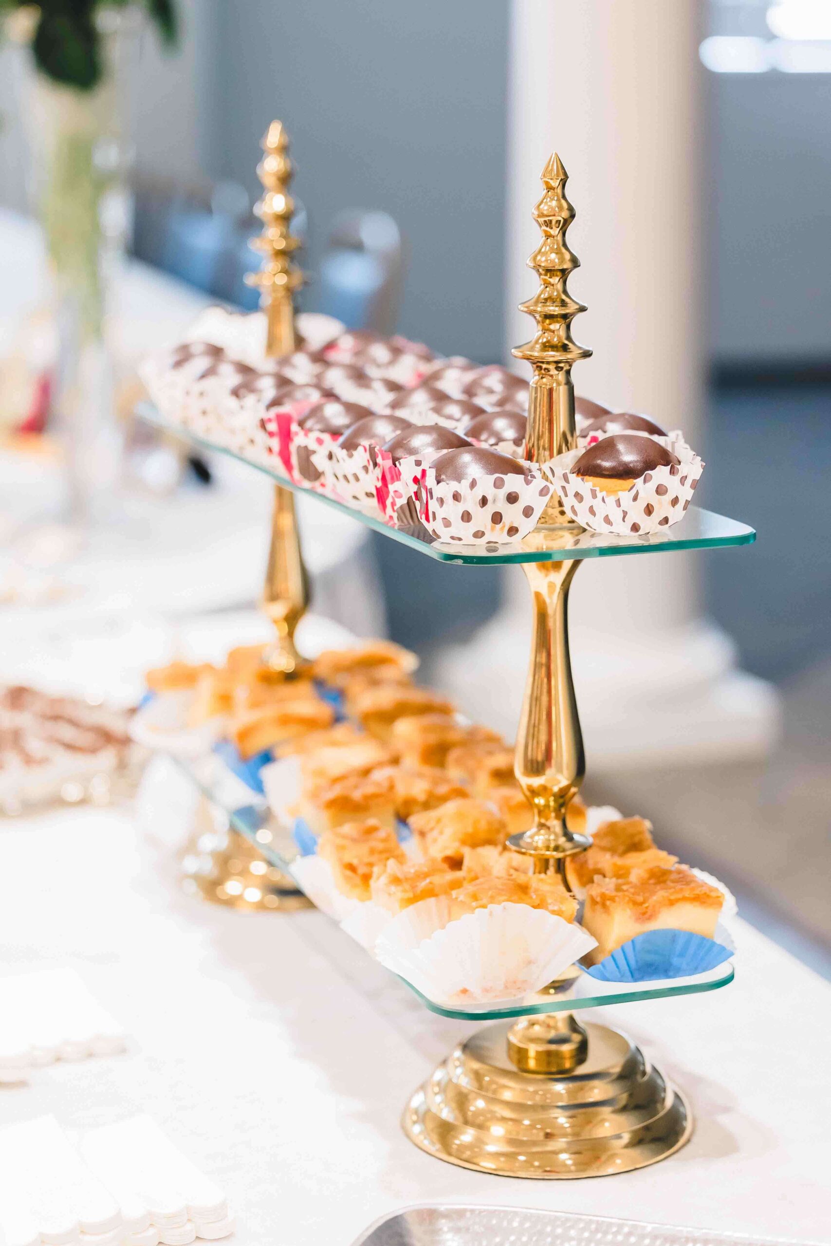 Close-up of a dessert table featuring two-tiered gold stands. The top tier holds chocolate-coated treats in decorative wrappers, and the bottom tier has golden-brown pastries in paper cups.