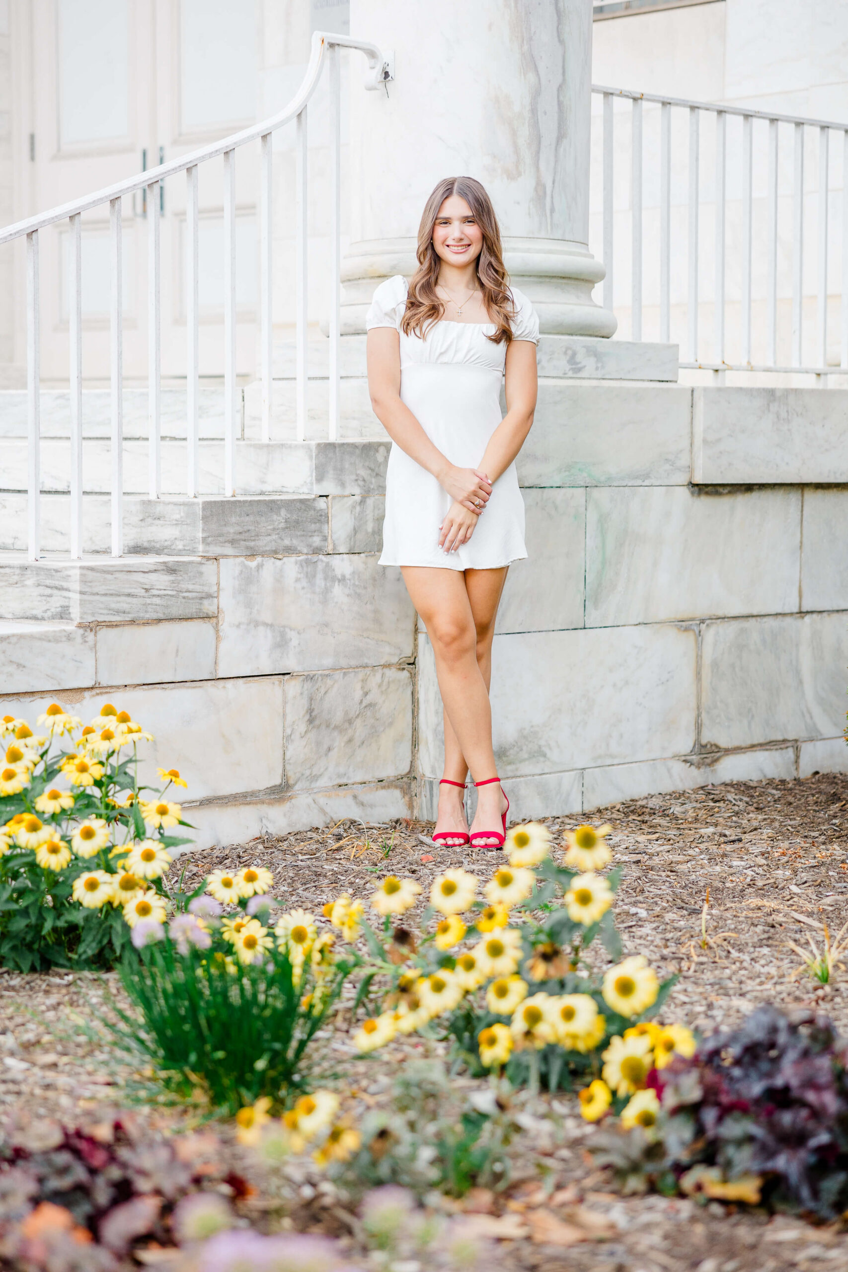 A woman in a white dress and red shoes stands in front of a marble structure with steps in a garden