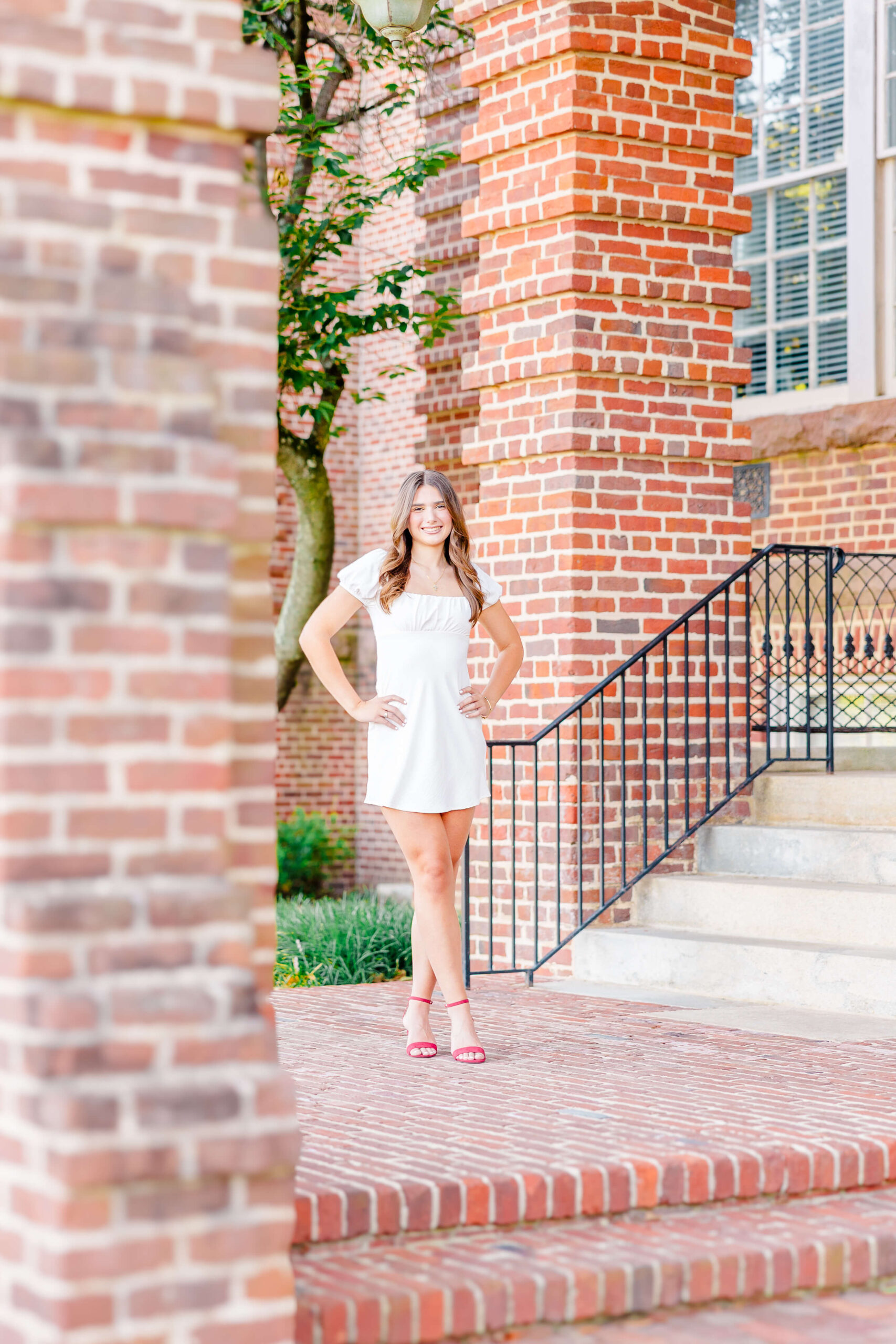 A graduate stands on a brick porch wearing a white dress and red heels with hands on her hips after visiting boutiques in fayetteville