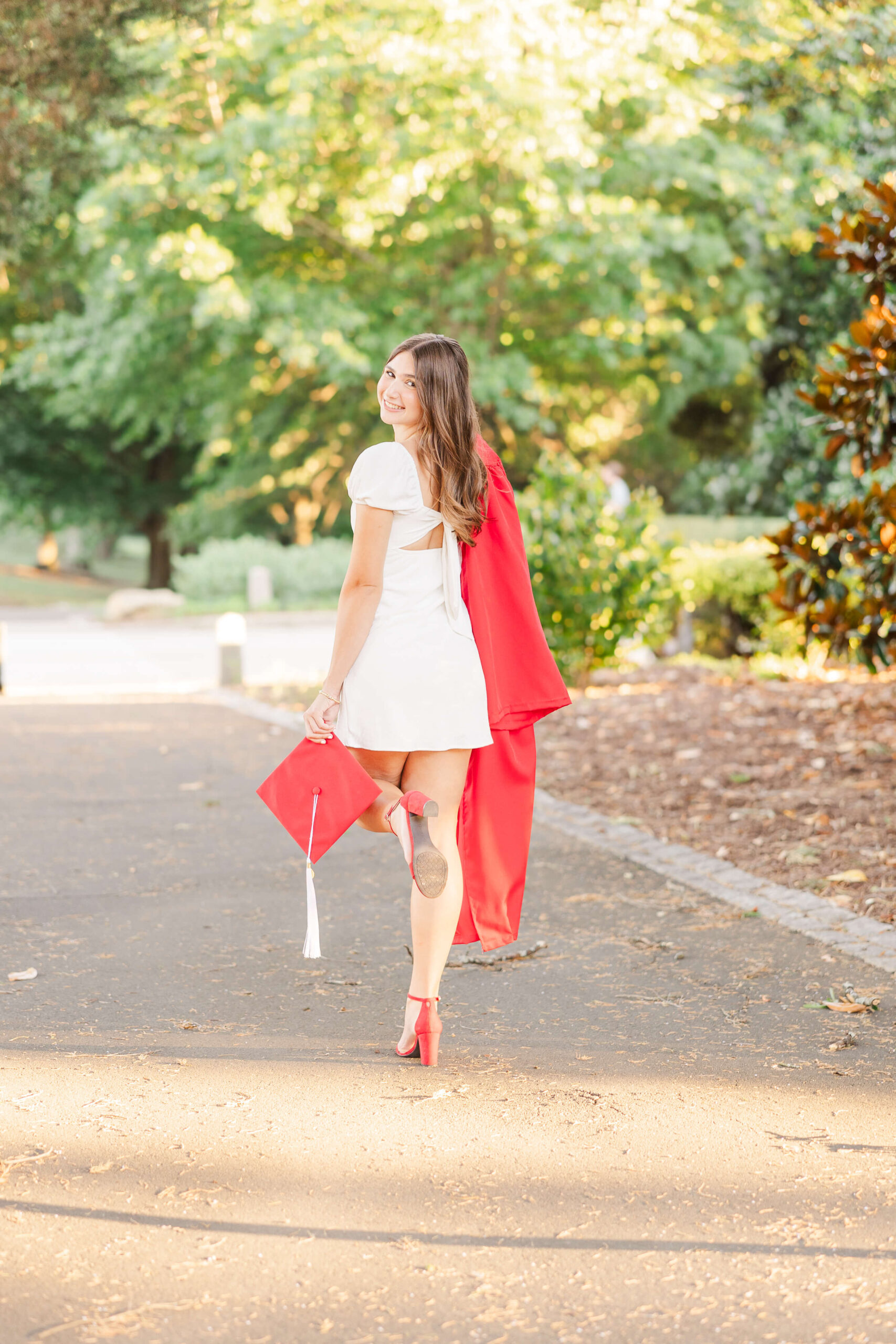 A graduate in a short white dress and red high heels walks away on a sunlit pathway, holding a red graduation cap in one hand and draping a matching gown over her shoulder, looking back and smiling after visiting boutiques in fayetteville