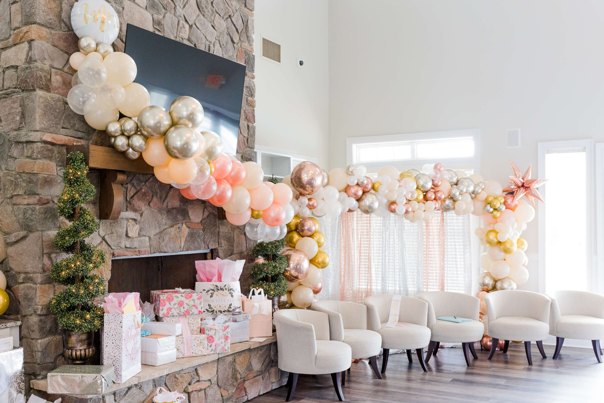 A decorated room features a stone fireplace adorned with colorful balloons in shades of white, gold, pink, and orange and presents