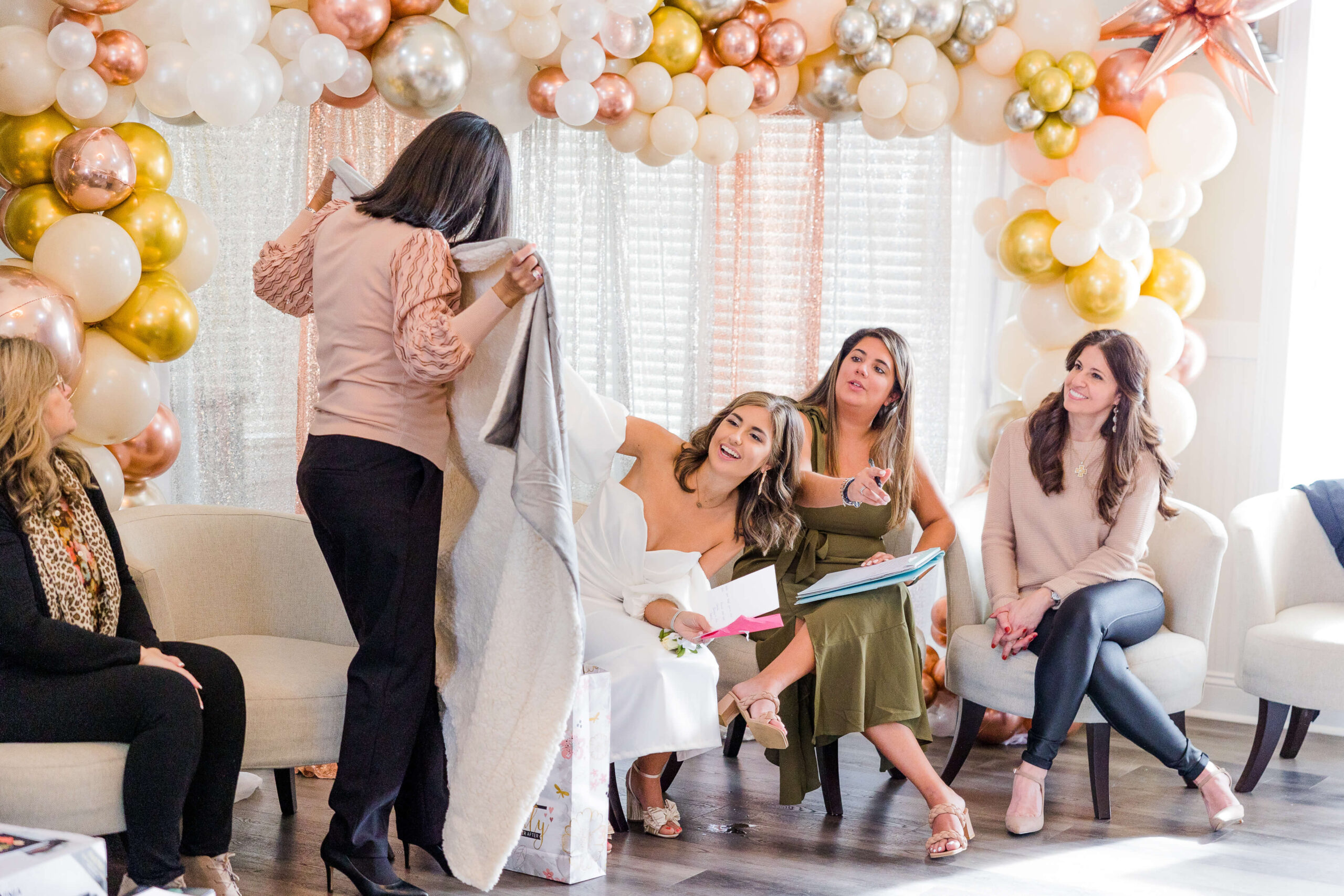 A group of four women sitting on white chairs, surrounded by a balloon arch in shades of gold, silver, and white while opening presents at one of the event venues in Fayetteville, NC