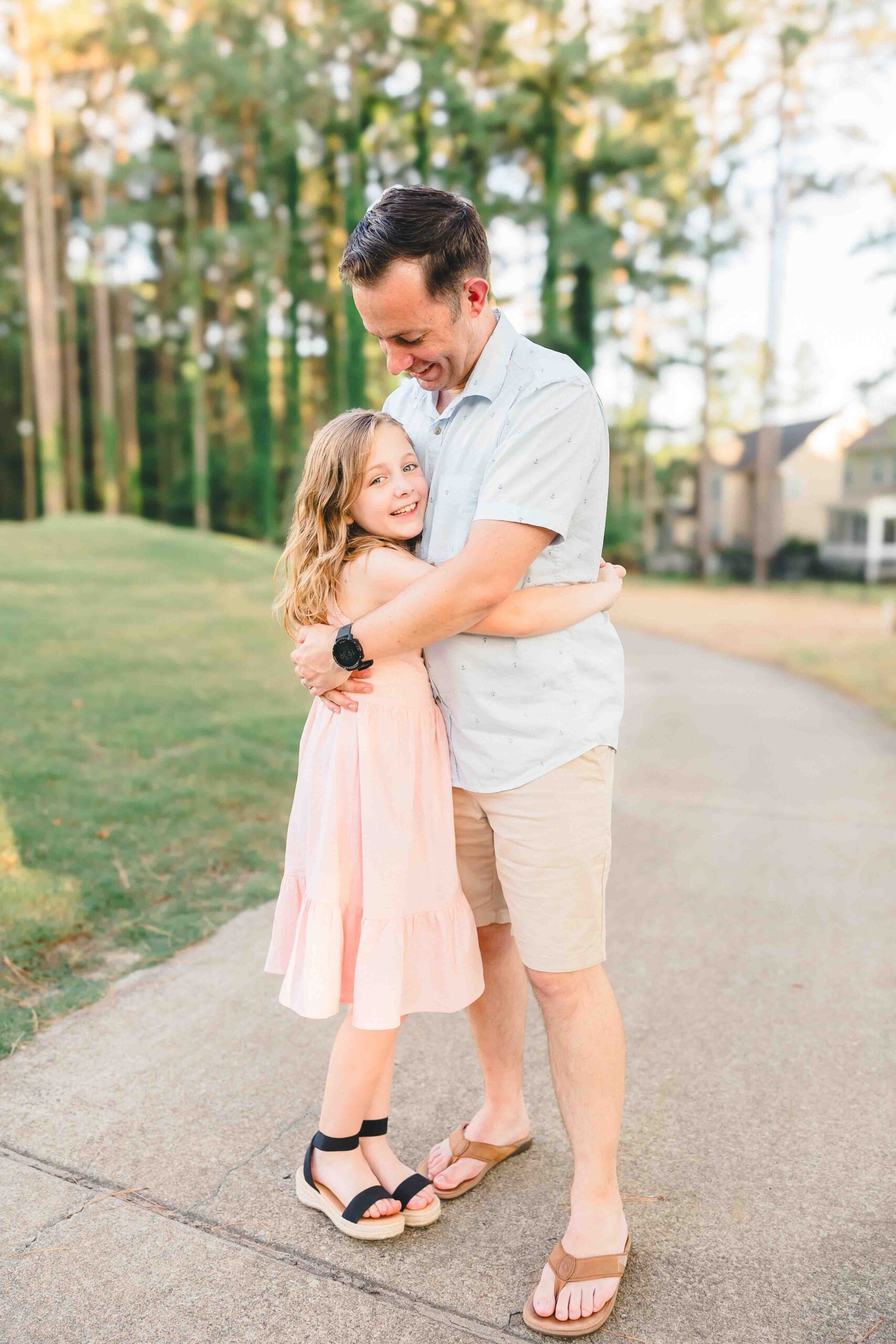 A young girl in a pink dress hugs dad while standing in a sidewalk at sunset after visiting pediatricians in Fayetteville