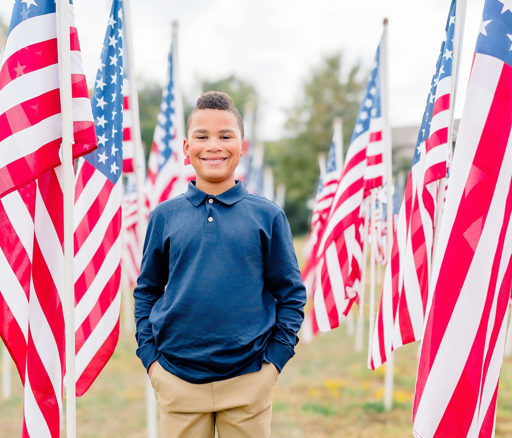 A young boy with a blue shirt and beige pants stands smiling in a field surrounded by American flags on poles.