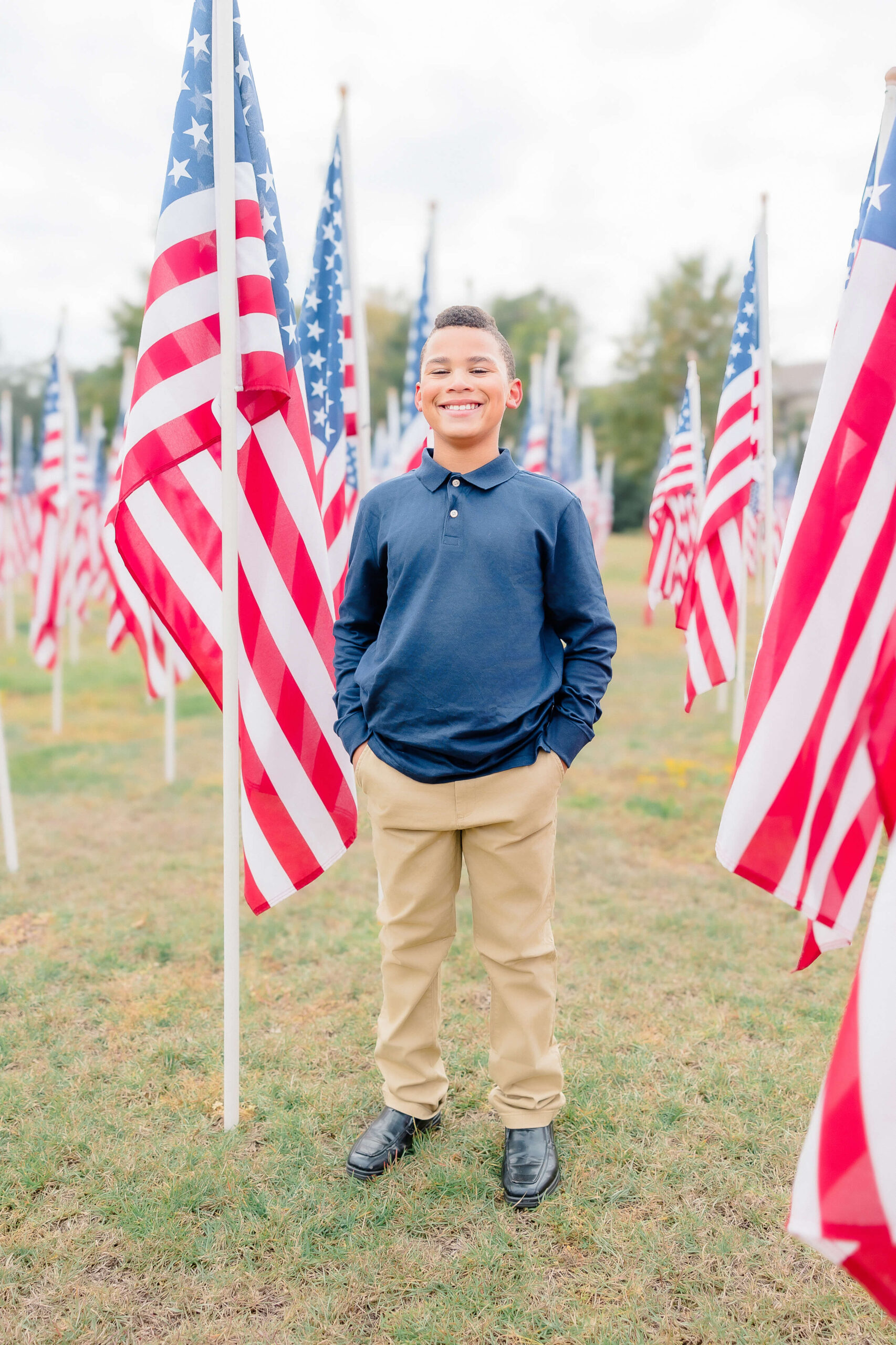 A young boy stands outdoors among numerous American flags displayed in rows in a navy shirt with hands in pockets after starting Youth sports leagues in Fayetteville NC