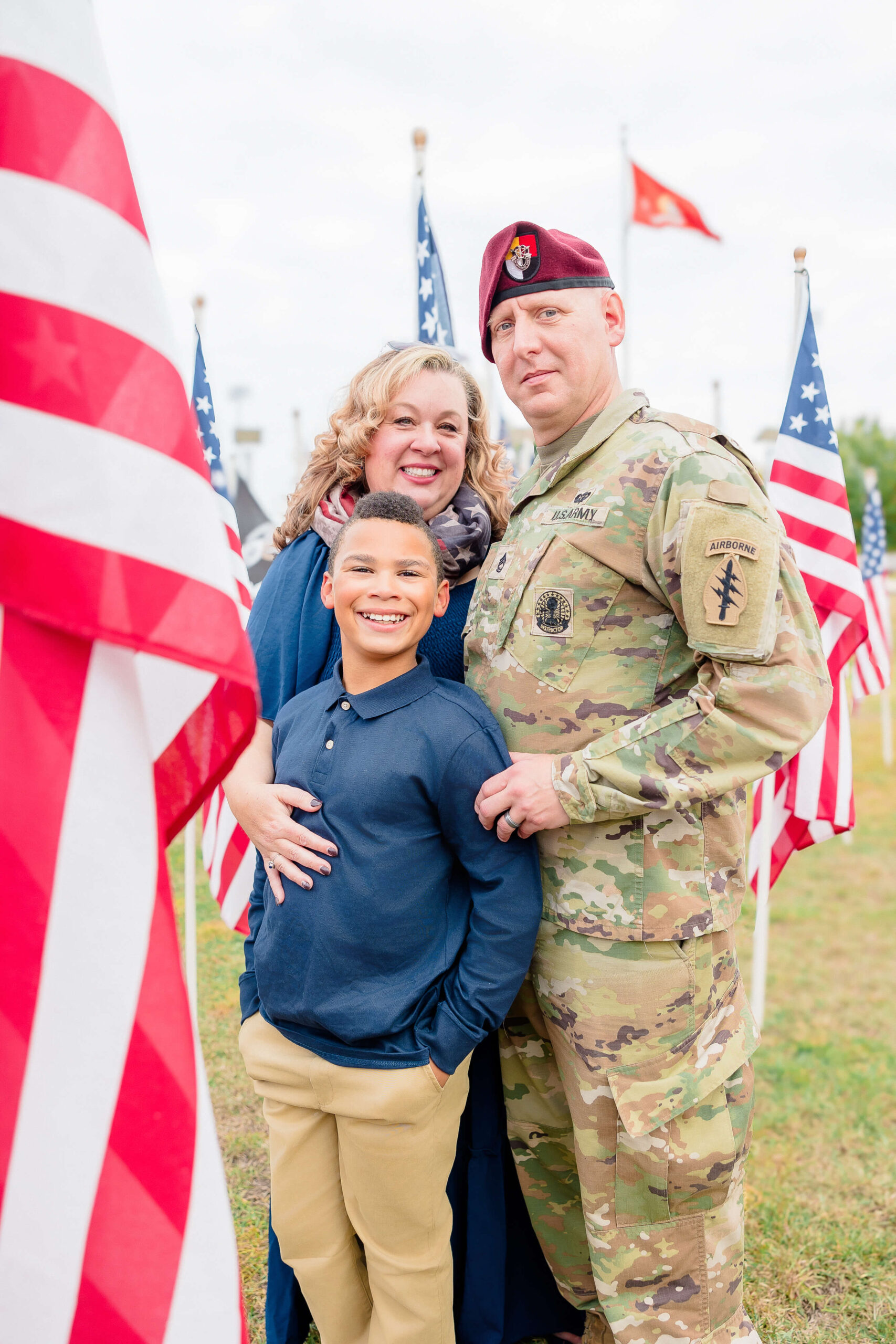 A father in a military uniform stands alongside a smiling child and wife in a field of American flags after exploring Youth sports leagues in Fayetteville NC