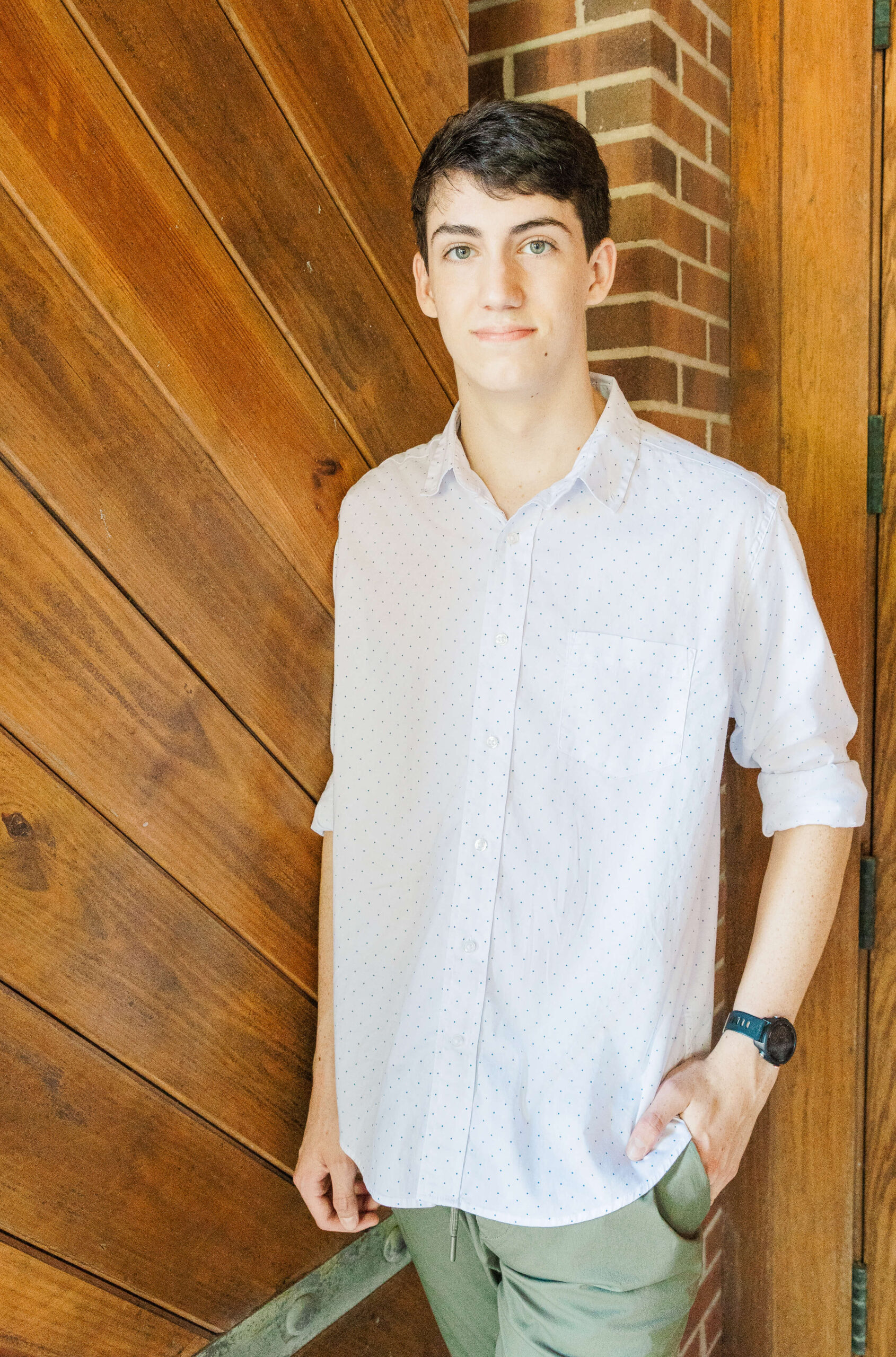 A young man in a white shirt and khaki pants stands against a wooden and brick wall