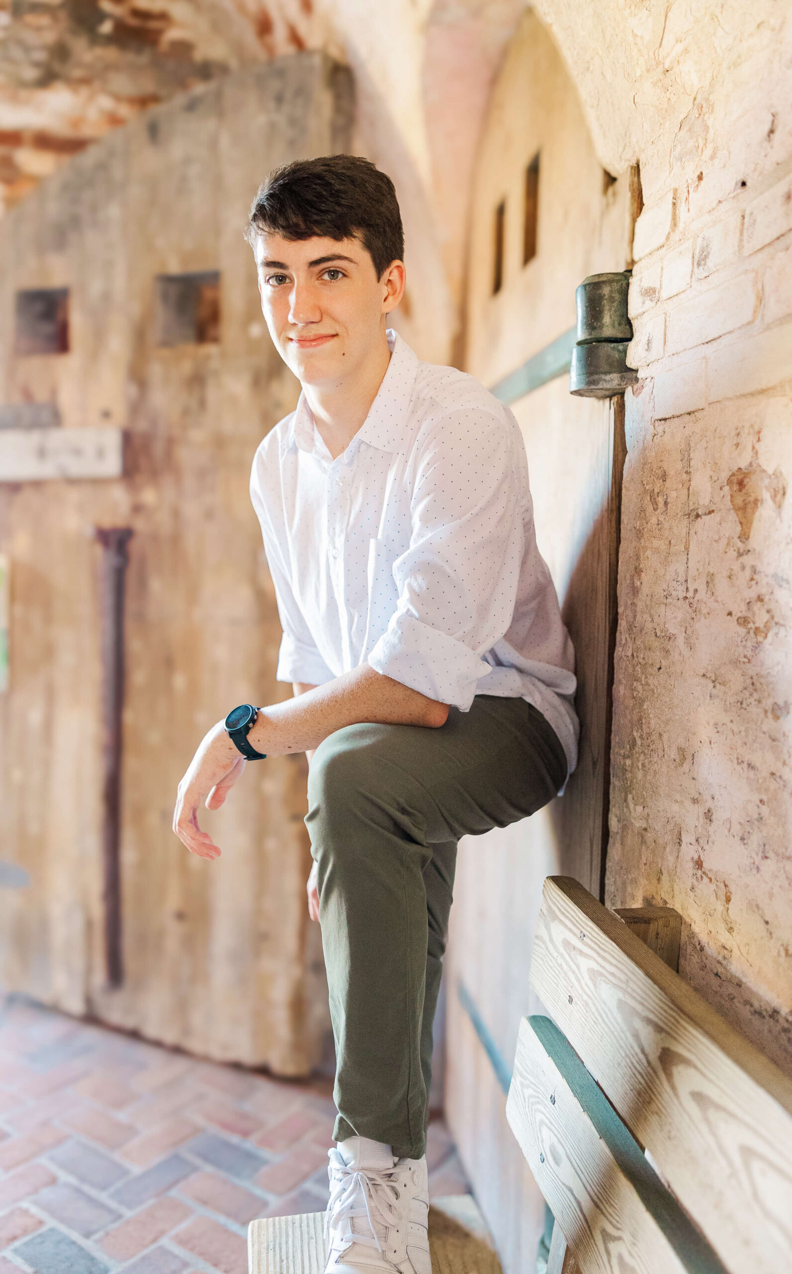 A young man with short dark hair, wearing a white shirt and olive pants, leans on a wooden bench in a rustic setting after visiting bowling alleys in Fayetteville nc