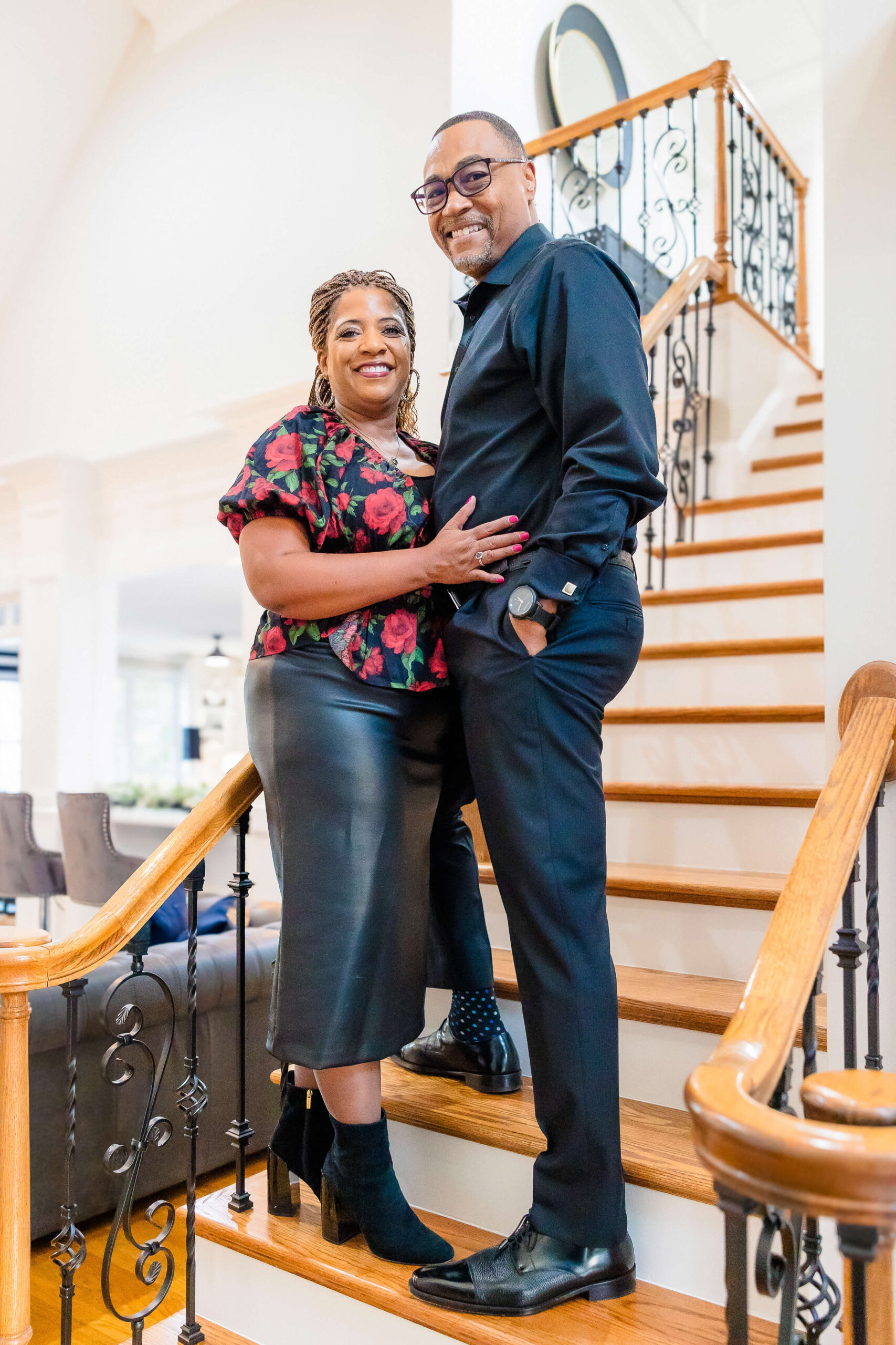 A smiling couple stands on a wooden staircase in their home in black and red after visiting movie theaters in Fayetteville NC