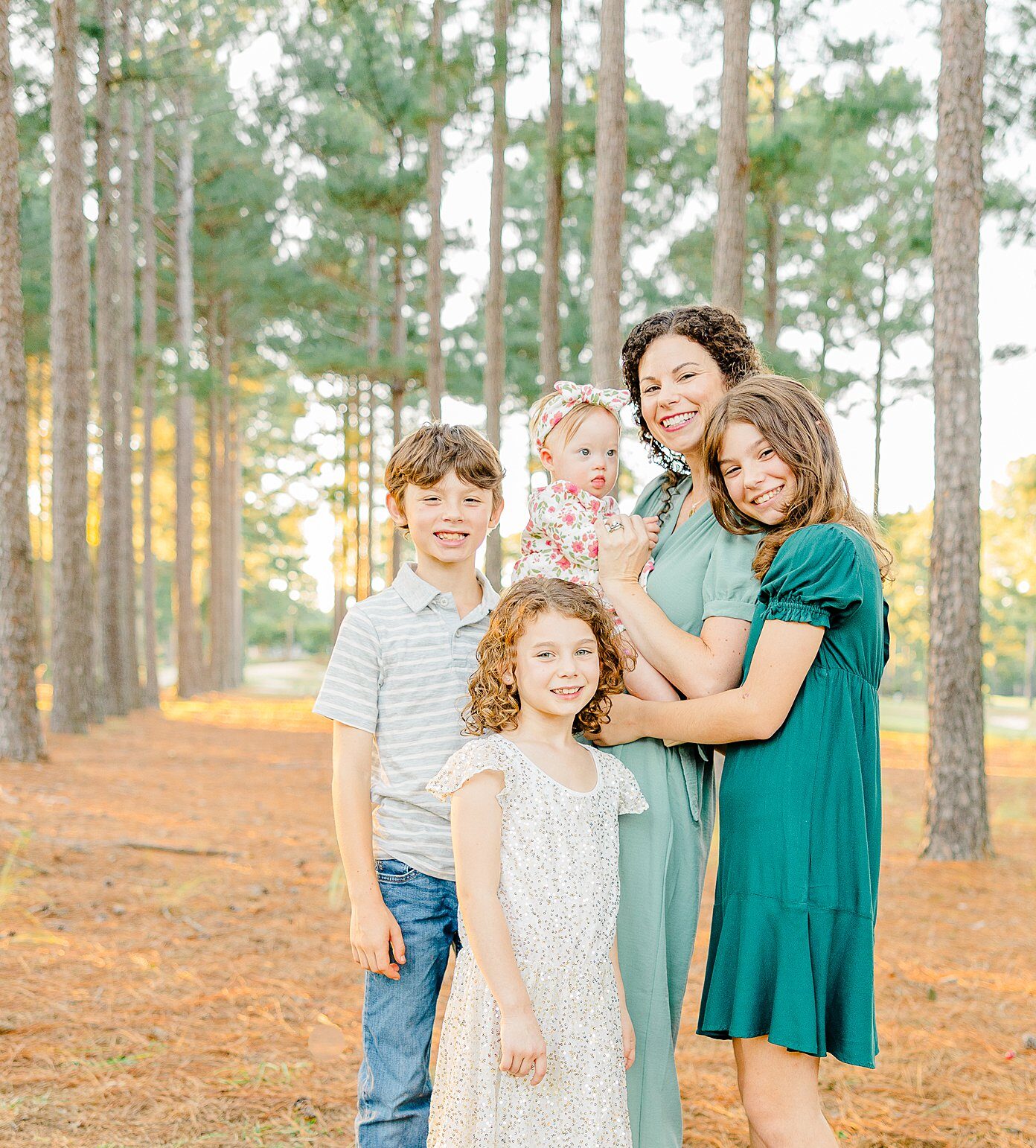 A happy mom stands with her three young children and holding a baby in a pine forest