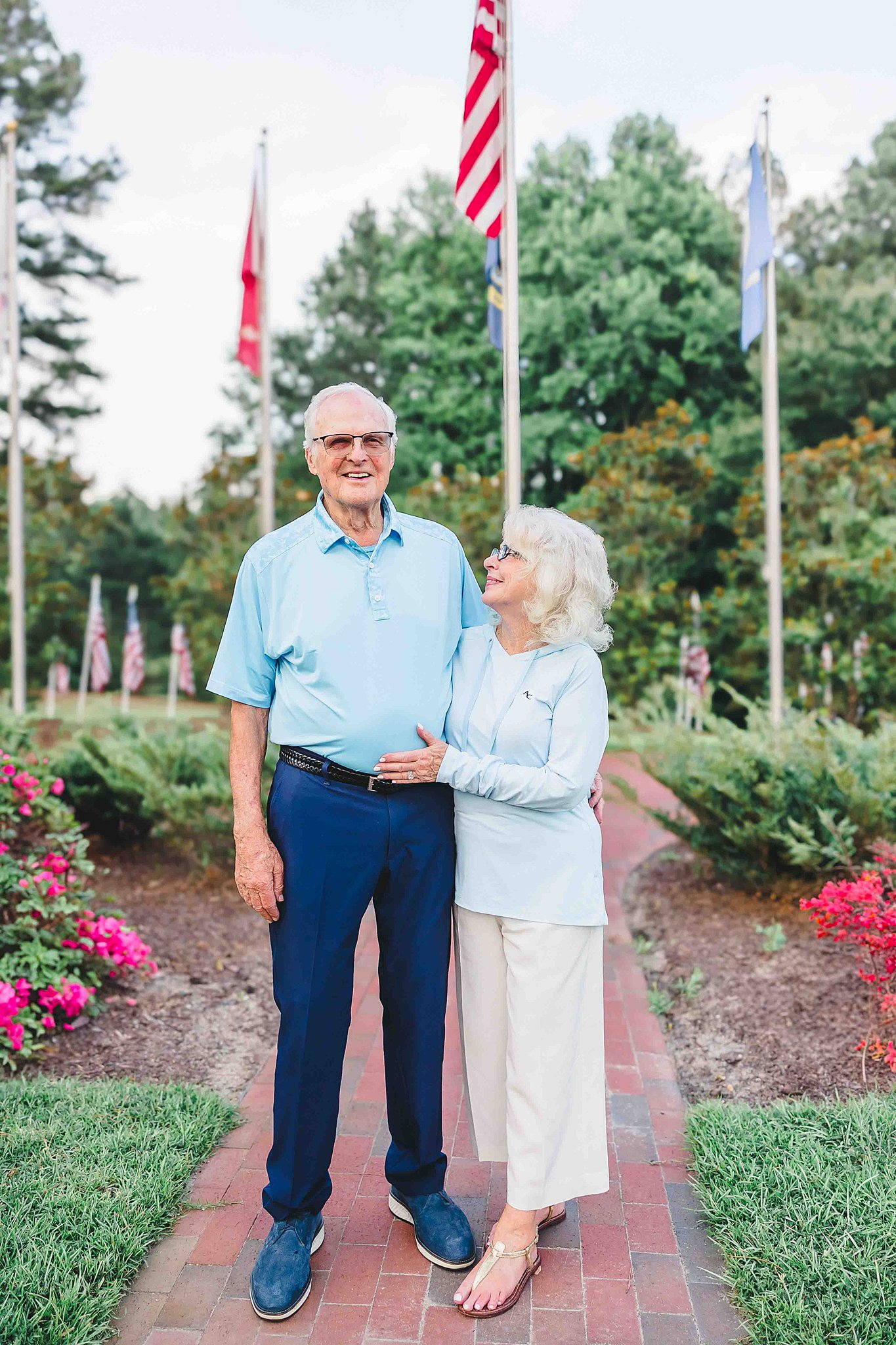 An elderly couple stands on a brick path lined with greenery, american flags and pink flowers.