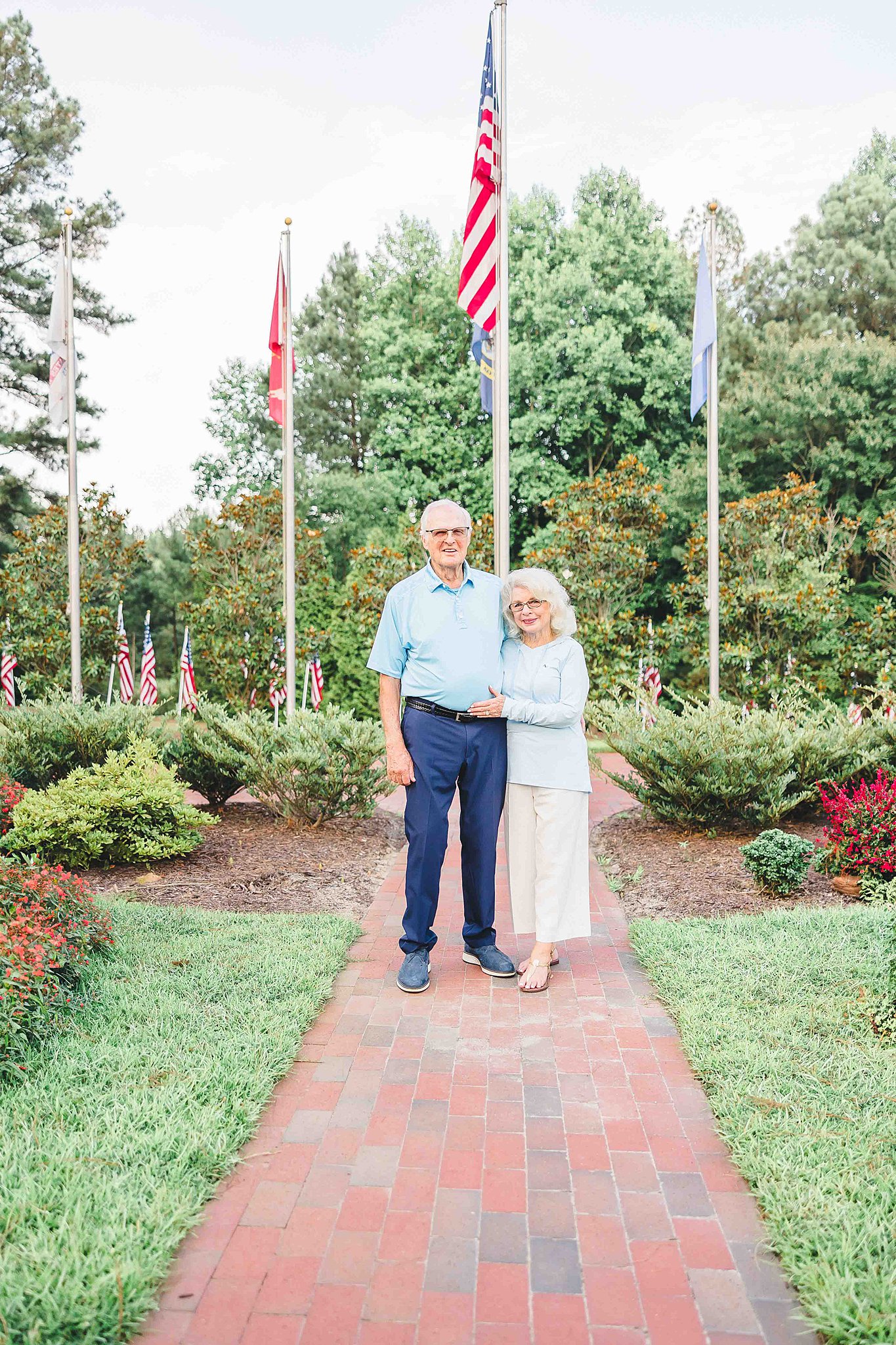 An elderly couple stands on a brick path in a park, surrounded by green shrubs and trees in front of many american flags after visiting Nail Salons in Fayetteville, NC