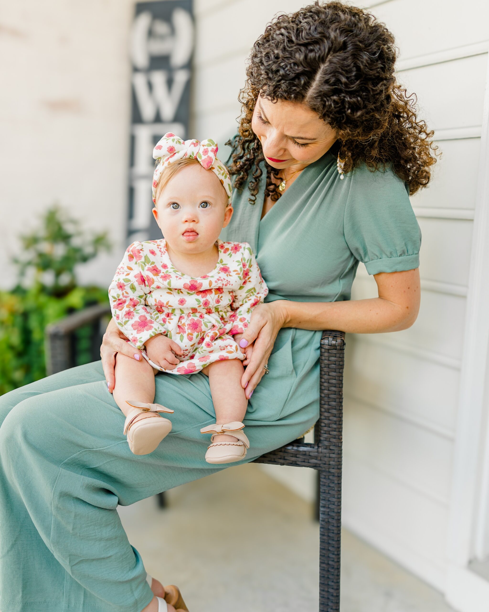 A mom with curly hair wearing a light green dress sits on a chair holding a baby on her lap.