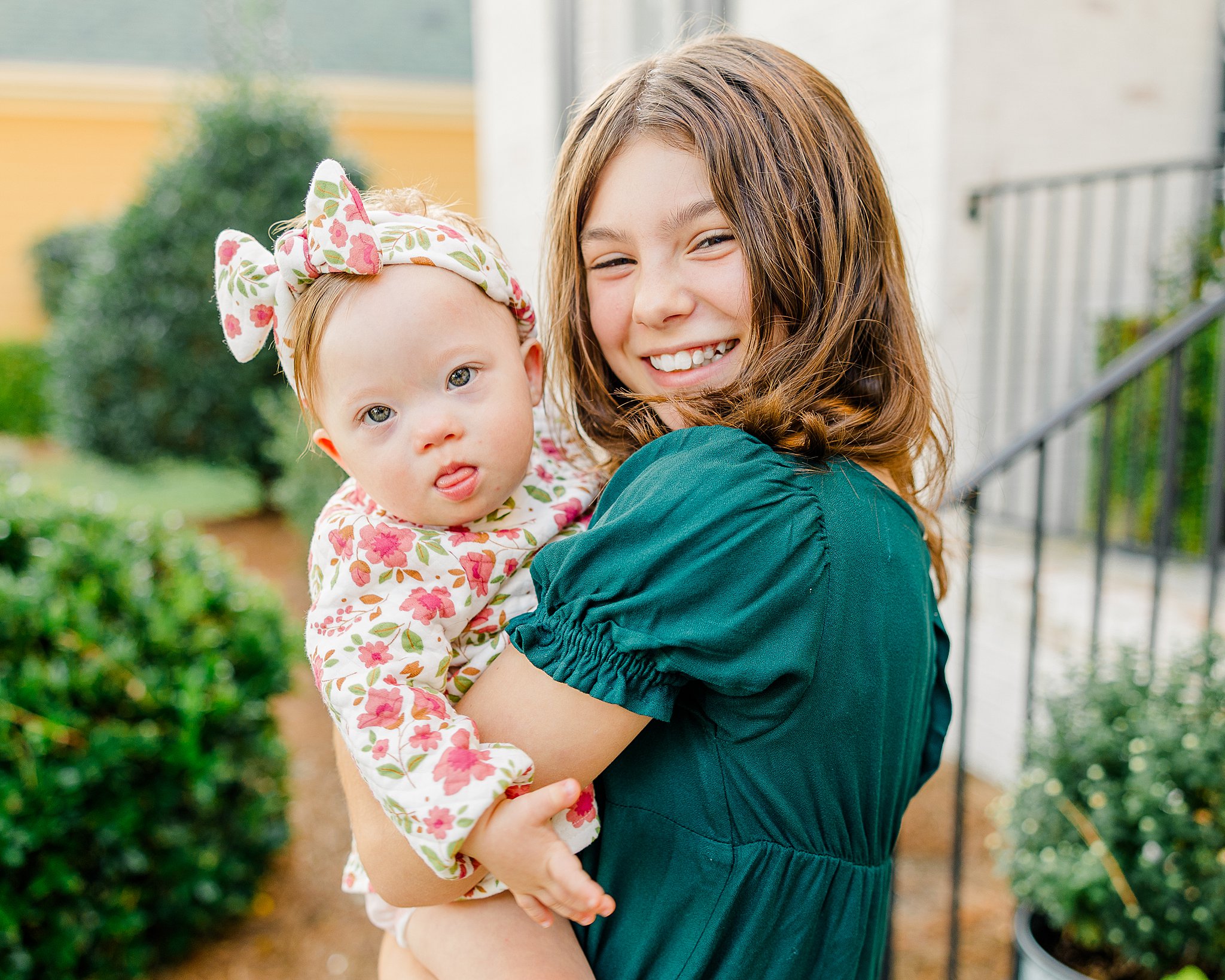 A young girl in a green dress holds her baby sister wearing a floral outfit and headband.