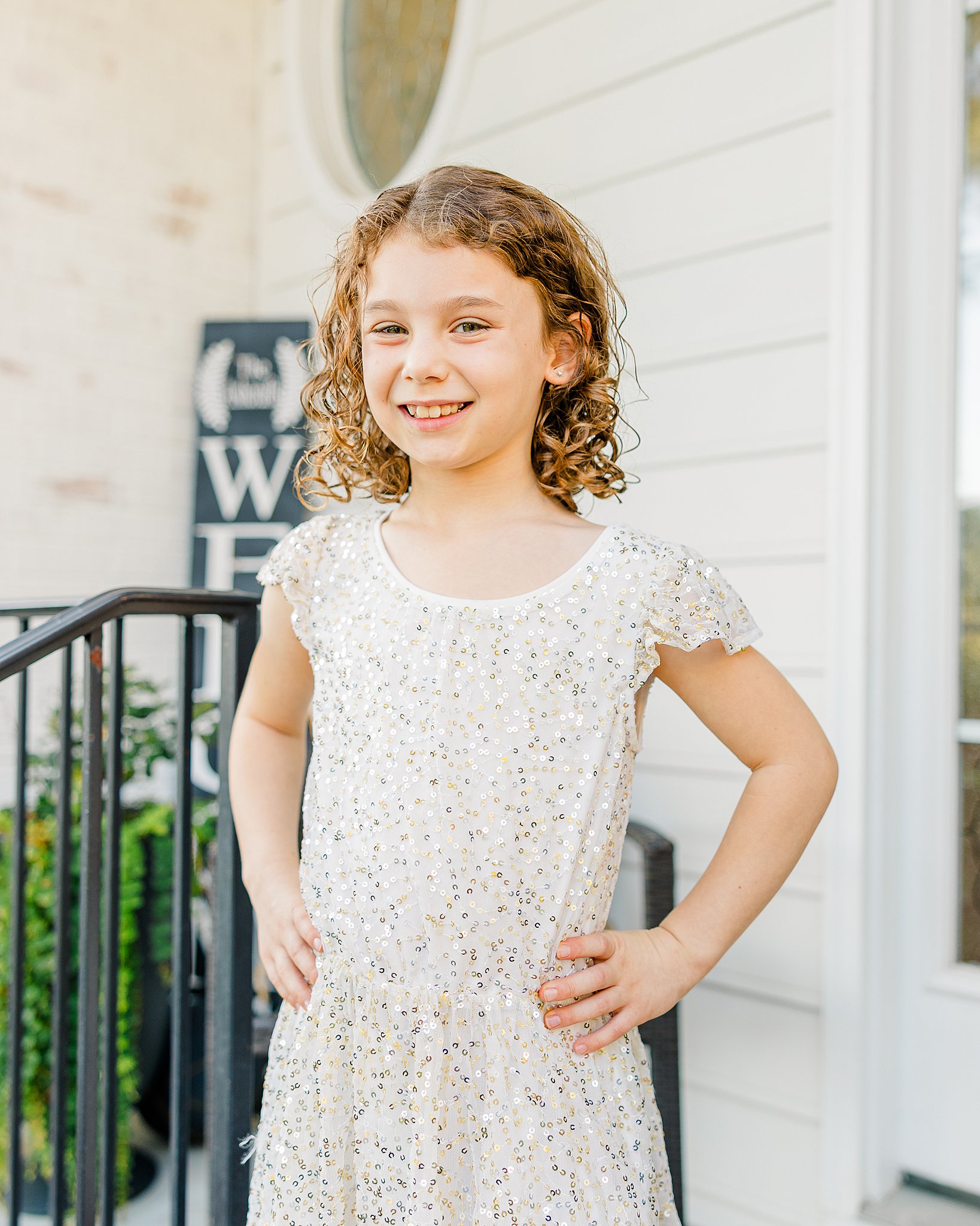 A young girl with curly hair smiles while standing on a porch in a white dress before visiting family friendly restaurants fayetteville, nc