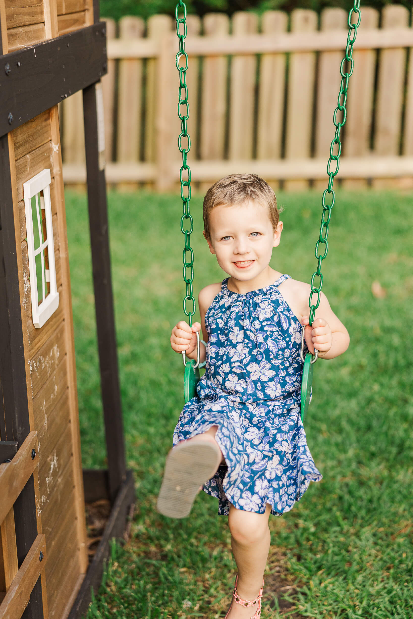 A young child in a blue floral dress is sitting on a swing attached to a wooden play structure.