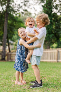 Three young siblings hugging and laughing in a grassy backyard as oldest boy carries baby before some summer camps in fayetteville