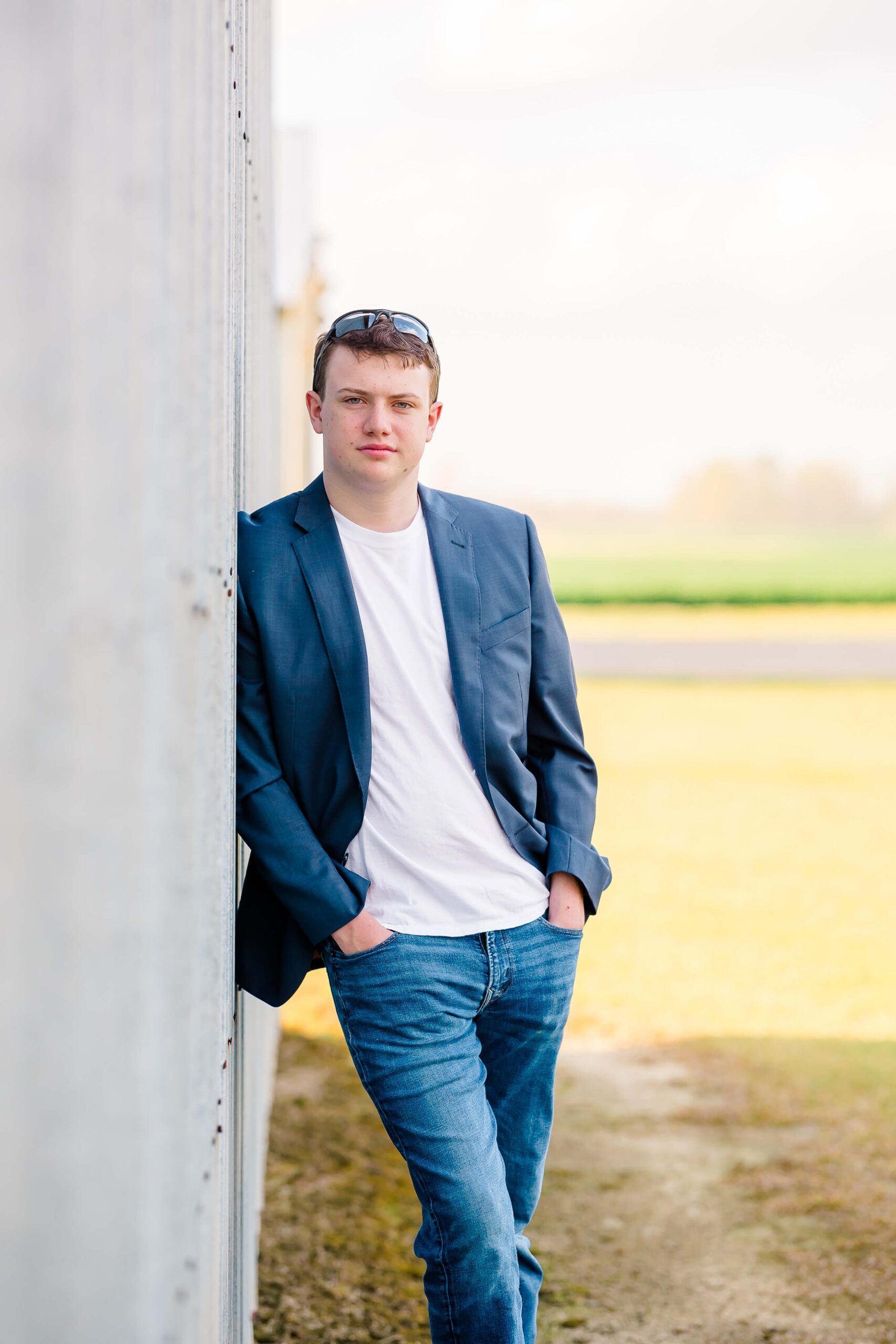 A young man wearing a blue blazer, white t-shirt, and jeans stands with hands in pockets, leaning against a wall outdoors.