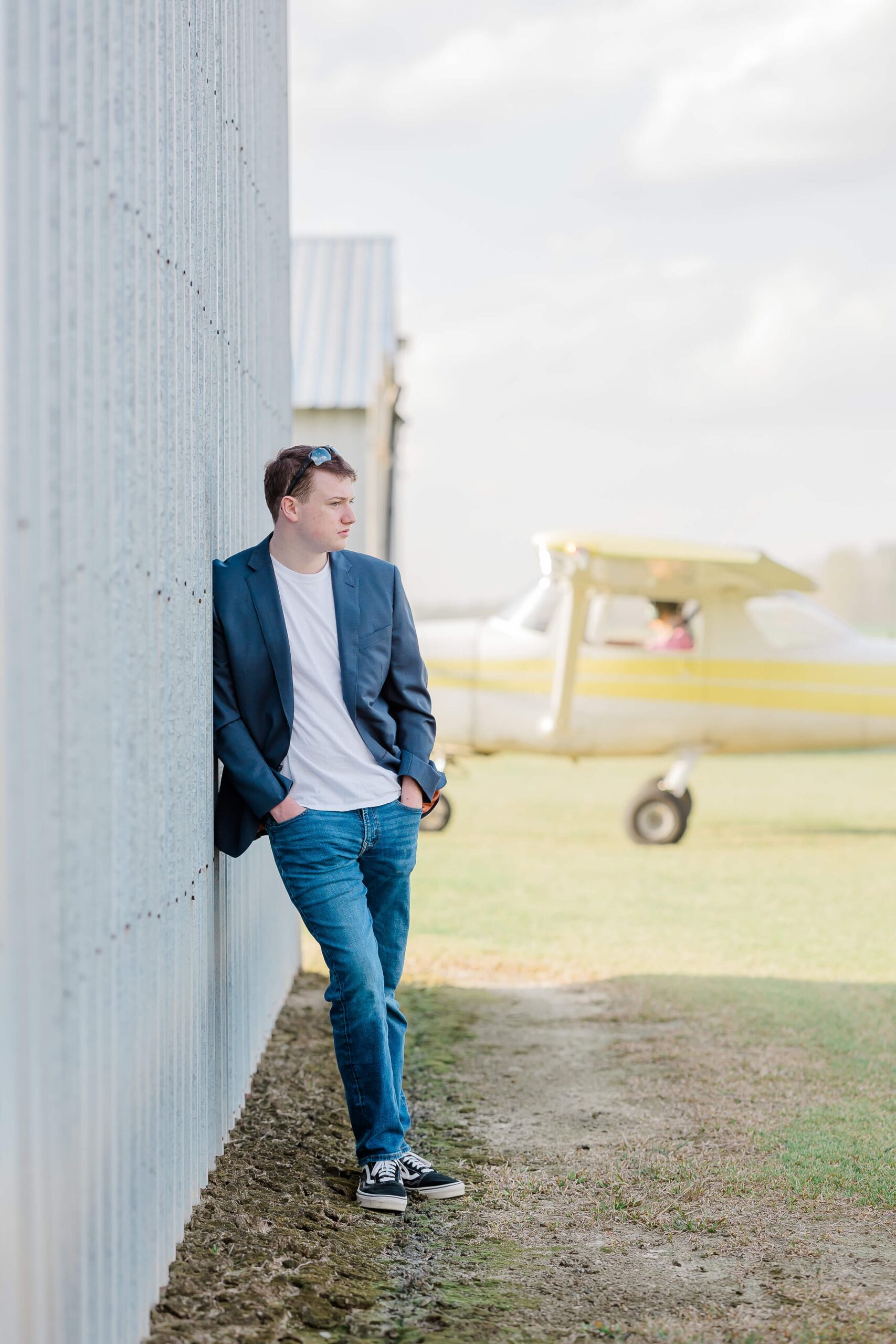 A man in a blue blazer, white shirt, and jeans leans against a corrugated metal wall in front of a small plane after finding tux rentals in fayetteville nc