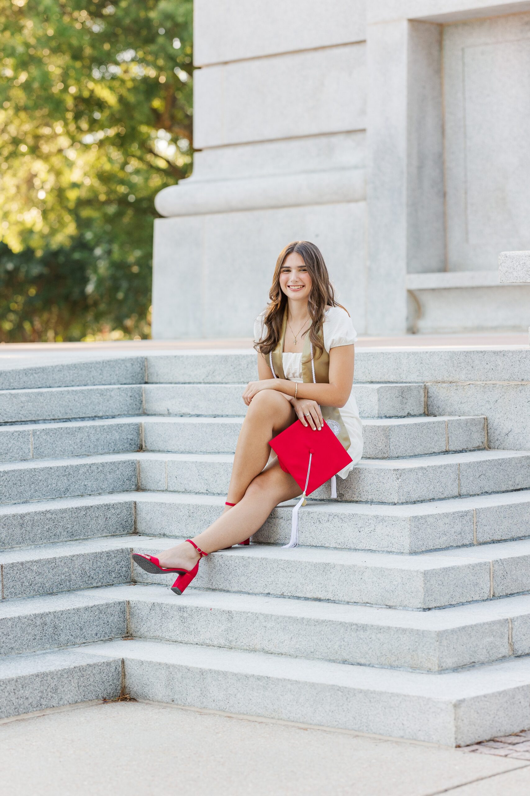 A graduate sits on stone steps of the NC State bell tower in a white dress and gold stole holding her red cap