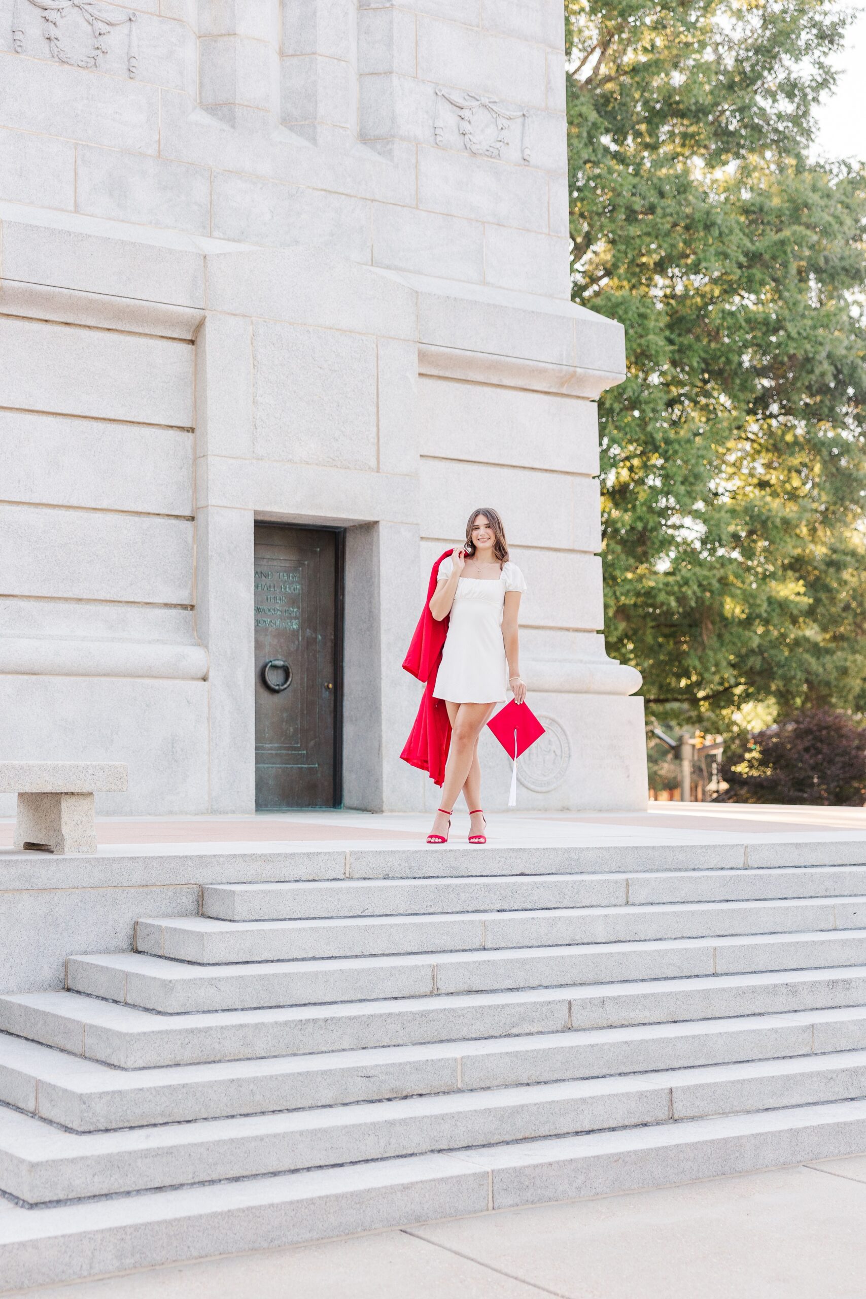 A college grad stands on stone steps outside the belltowers, holding a red gown over one shoulder at one of the colleges near fayetteville nc