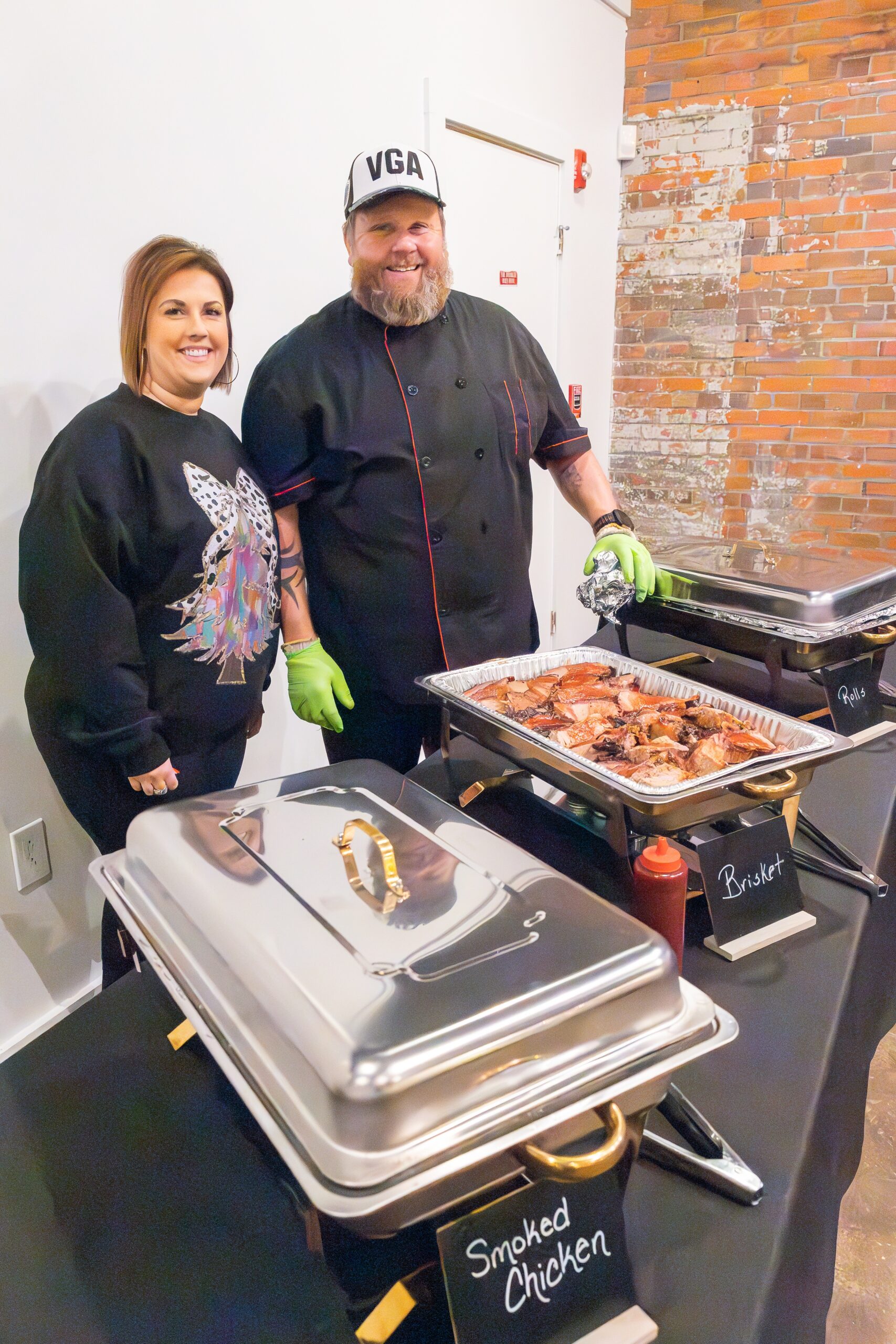 Two people stand behind a catering setup filled with BBQ.