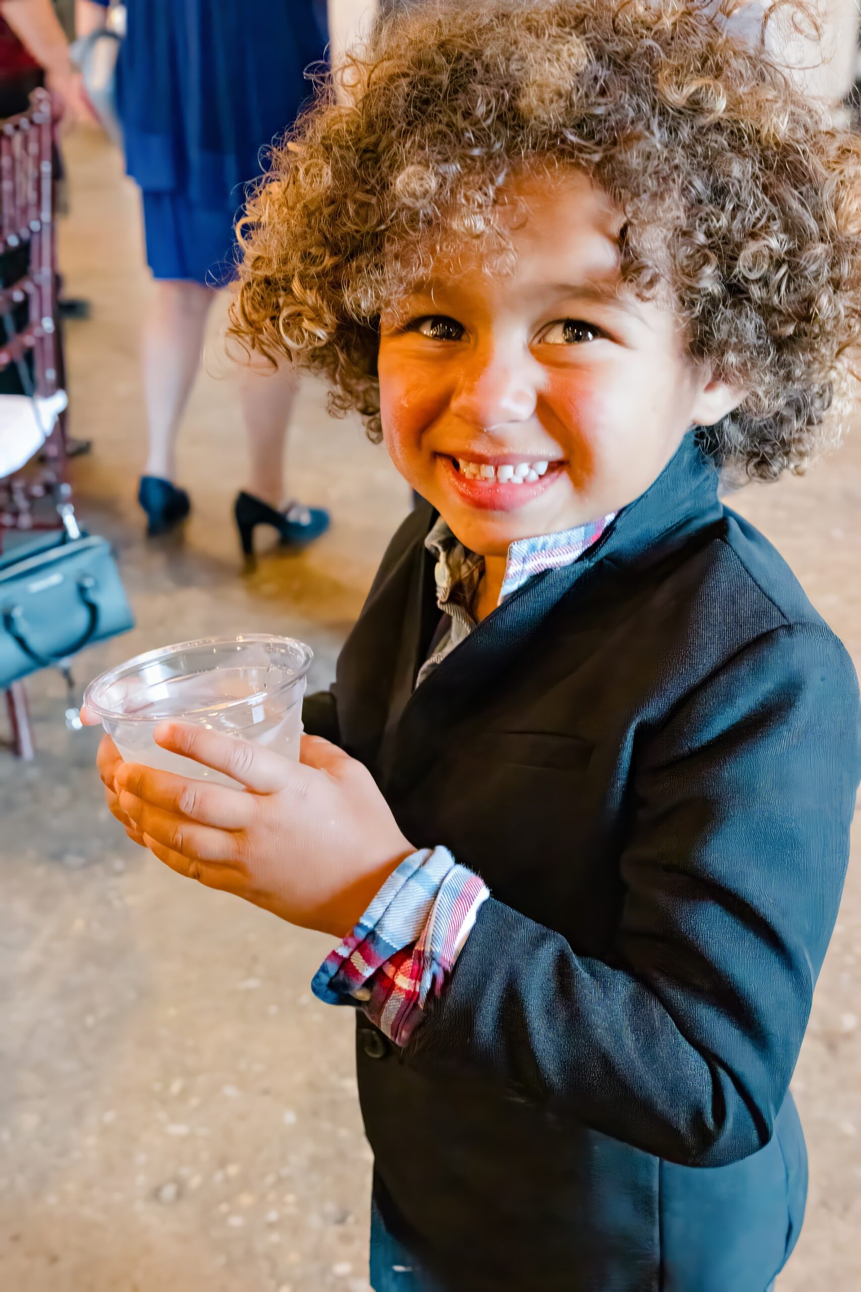 A young child with curly hair smiles while holding a plastic cup in a black suit jacket while enjoying event catering in Fayetteville, NC