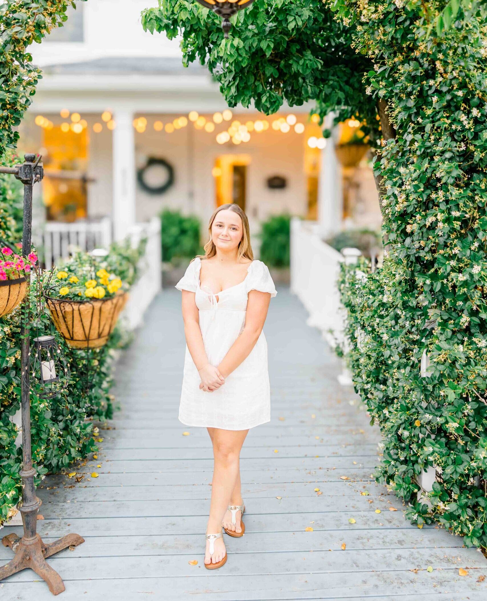 A woman in a white dress stands on a wooden path surrounded by greenery and hanging lanterns.