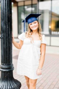 A graduate wearing a blue graduation cap and a white dress leaning against a black lamppost thanks to NC countdown to college.