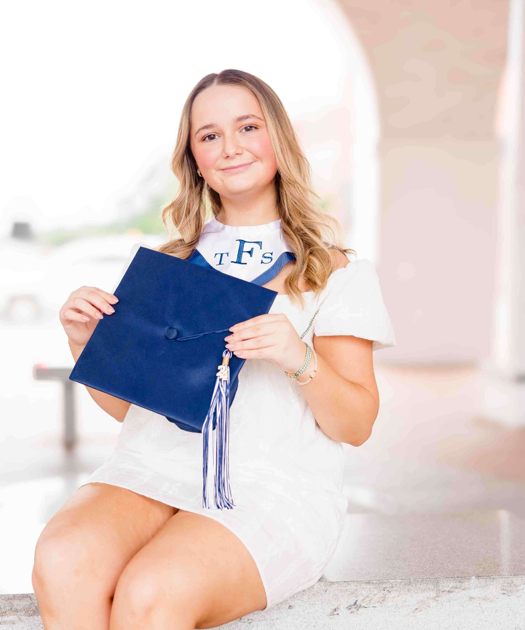 A graduate in a white dress sits on a stone step, holding a blue graduation cap with a tassel thanks to NC countdown to college.