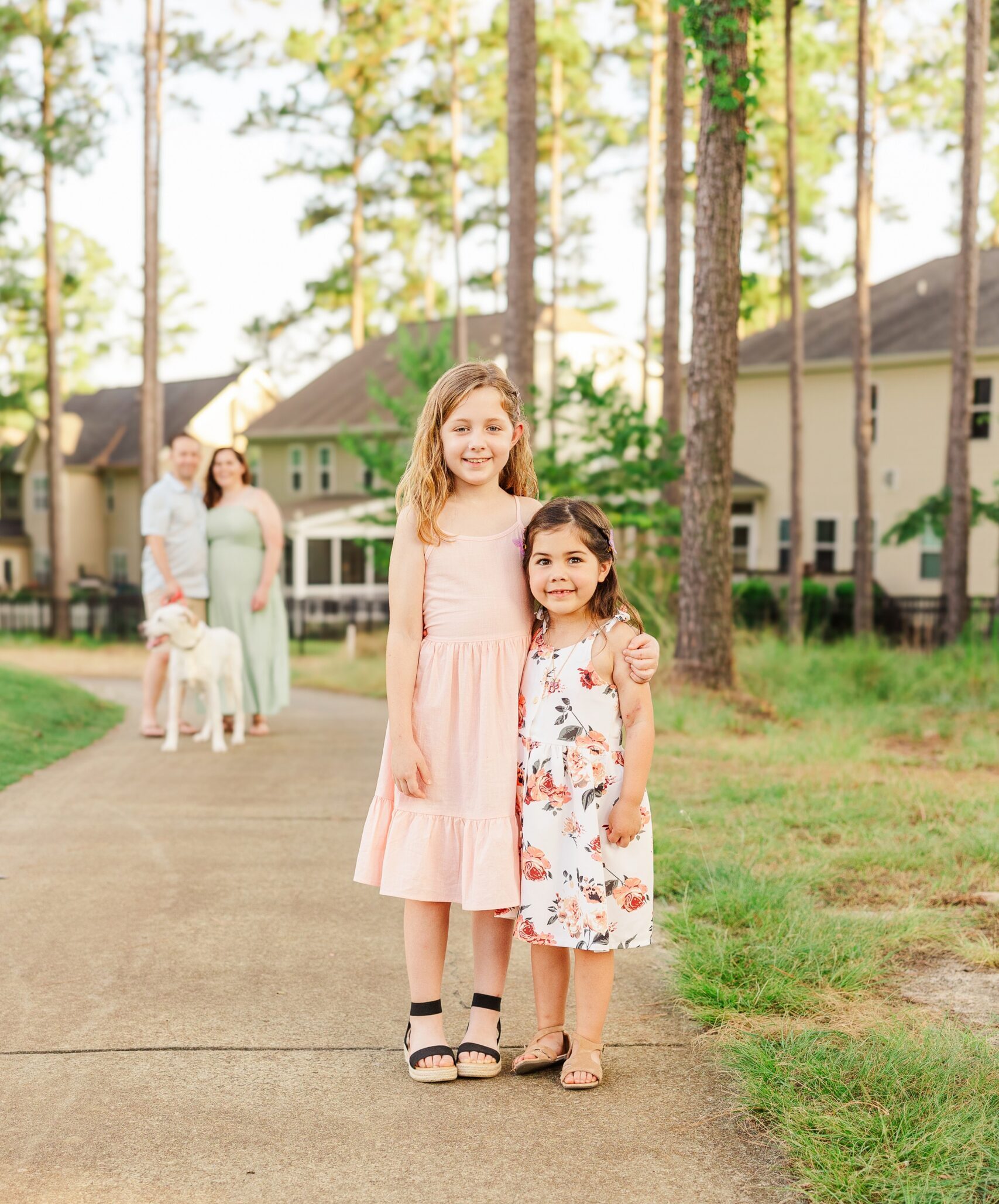 Two young sisters stand on a sidewalk, smiling in dresses as mom and dad look on