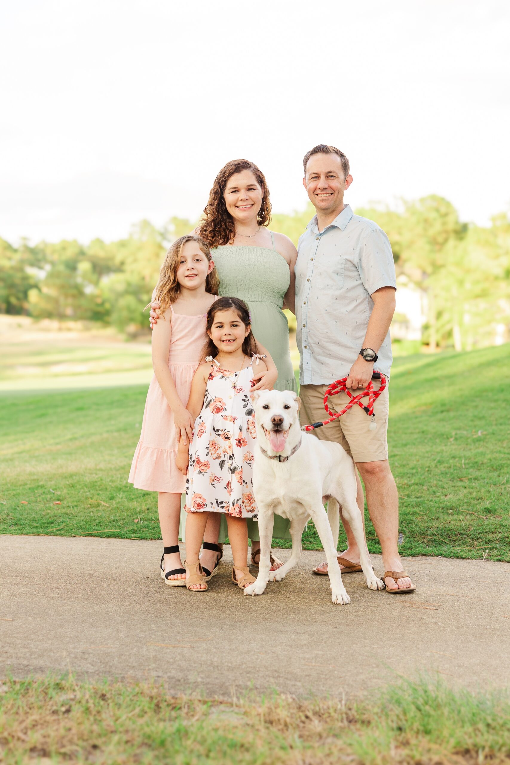 A family of four and a dog stand outdoors on a paved path with grass and trees in the background after visiting pediatric dentists in Fayetteville, NC