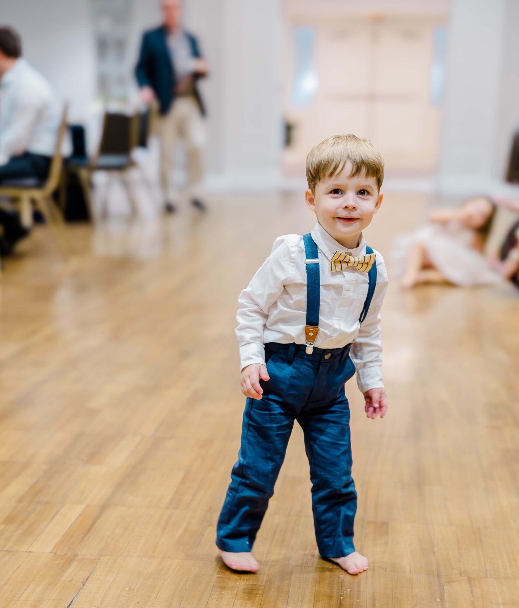 A toddler boy stands barefoot on a wooden floor, wearing a white shirt with a bow tie, blue suspenders, and blue pants at one of the small event venues fayetteville nc