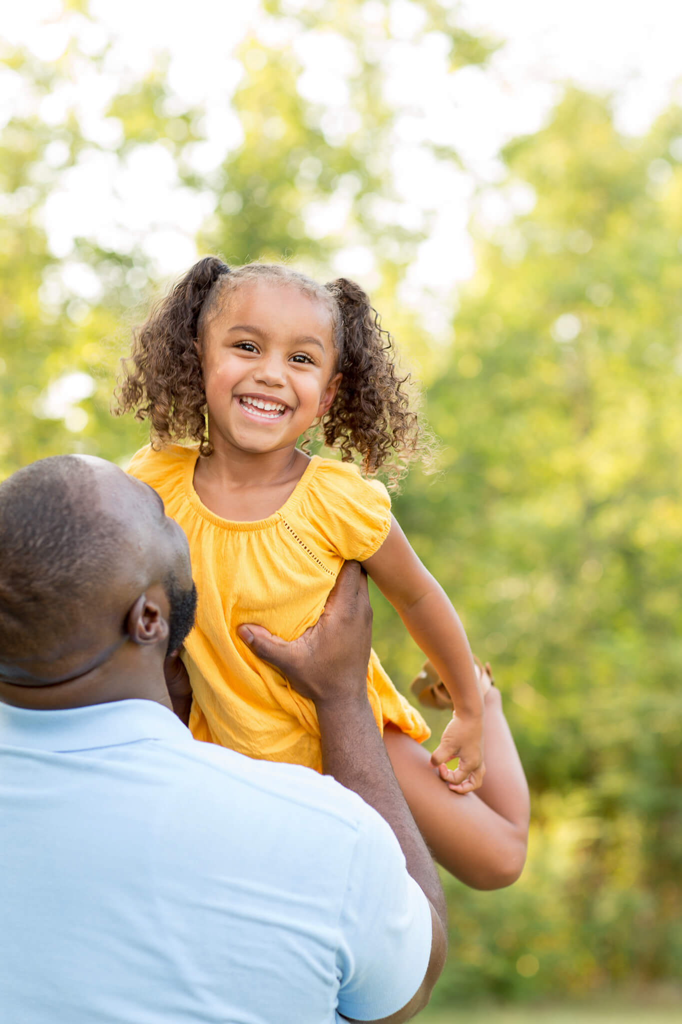 A dad in a light blue shirt lifts his smiling daughter in a yellow dress in a park after visiting toy stores in Fayetteville, NC