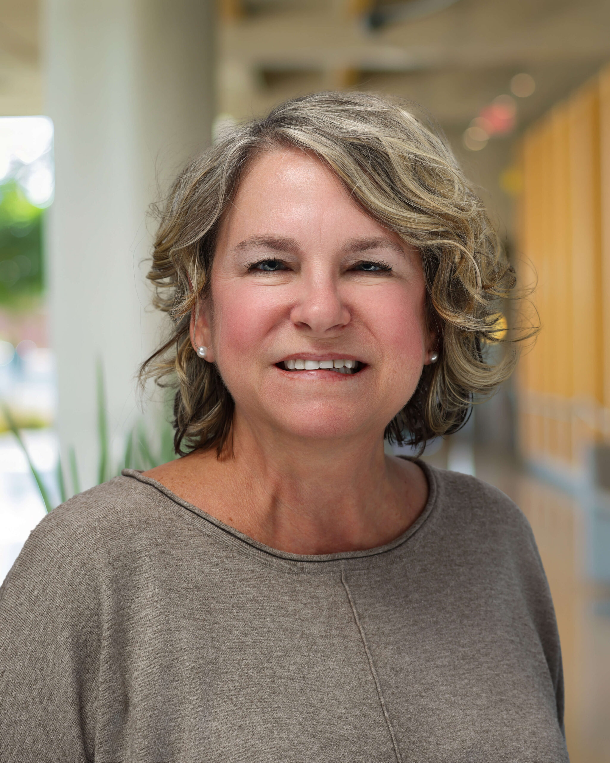A middle-aged woman with wavy, shoulder-length hair and a beige top smiles while standing indoors in a brightly lit hallway with columns and yellow accents in the background.
