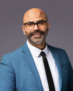 A man with a bald head, glasses, and a mustache with a beard is wearing a blue suit jacket, white shirt, and black tie. He is standing against a plain gray background and looking at the camera. Professional headshot photographer in Fayetteville NC creating corporate portraits for business professionals