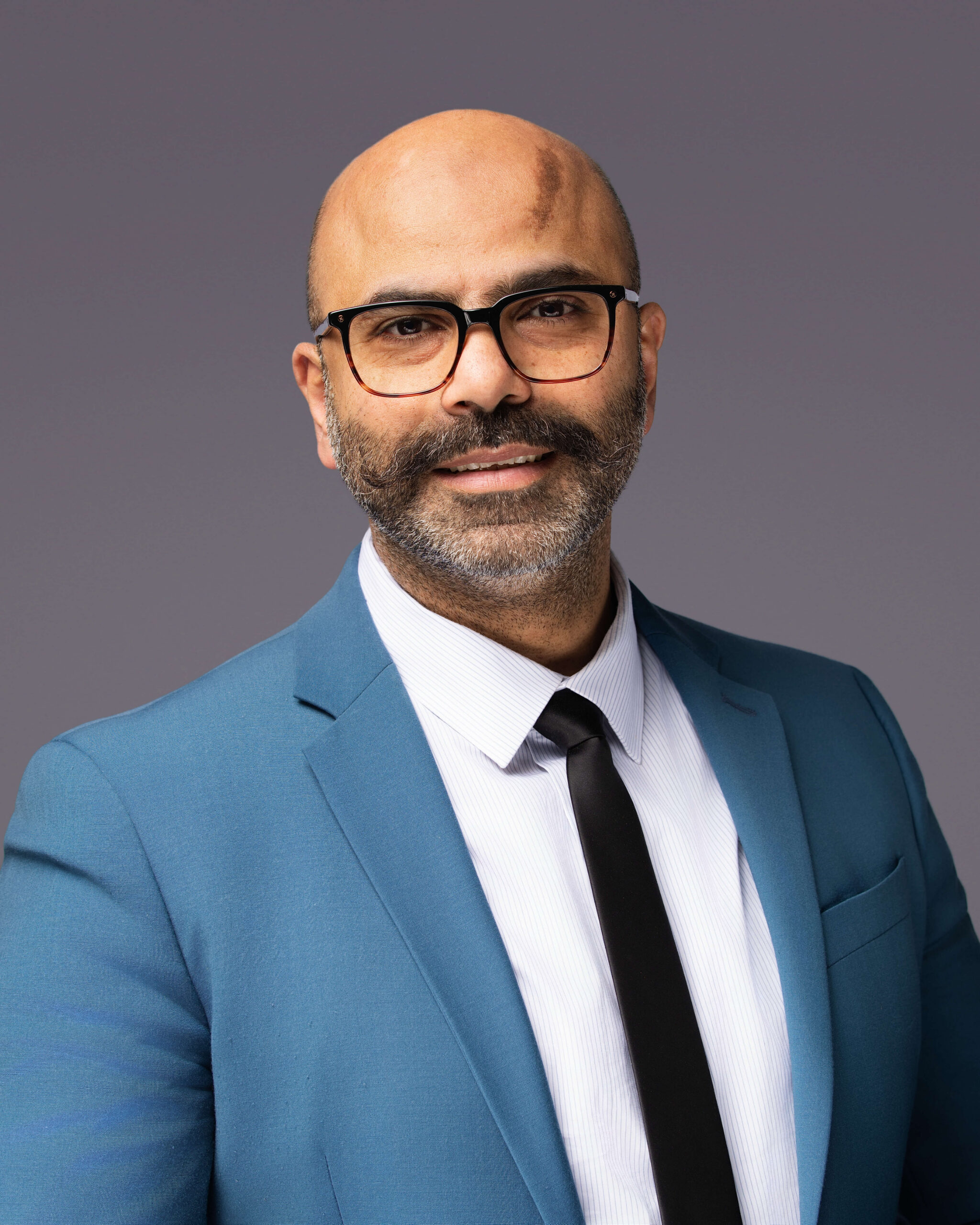 A man with a bald head, glasses, and a mustache with a beard is wearing a blue suit jacket, white shirt, and black tie. He is standing against a plain gray background and looking at the camera. Professional headshot photographer in Fayetteville NC creating corporate portraits for business professionals