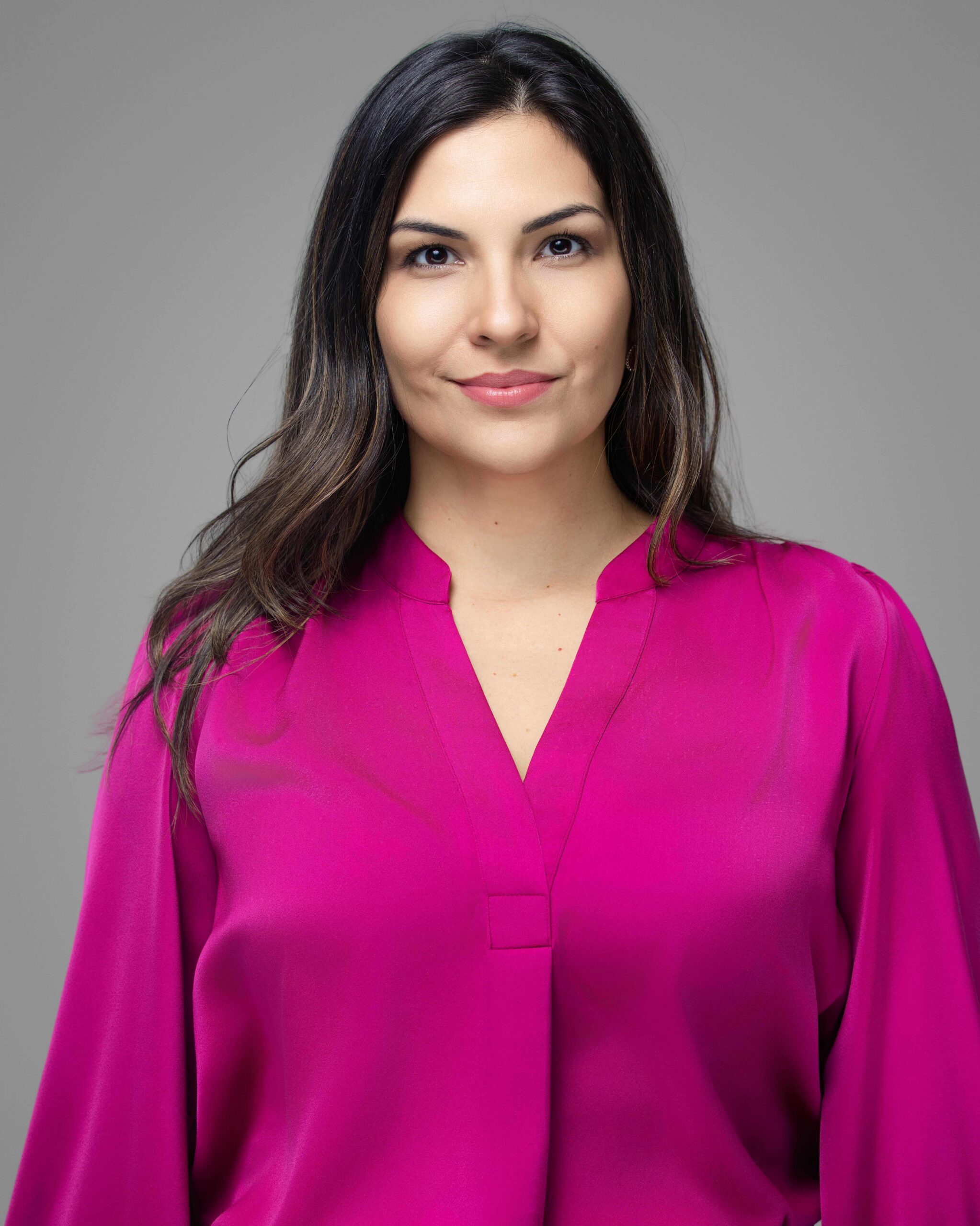 A woman with long brown hair wearing a bright pink blouse stands in front of a plain gray background, looking directly at the camera with a neutral expression.