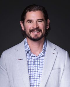 A man with medium-length brown hair and a beard is smiling. He is wearing a light gray blazer with a plaid shirt and a small lapel pin that reads "SAPMIX" on the left lapel. The background is dark gray.