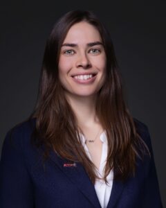 A woman with long brown hair, wearing a dark blazer over a white shirt, stands in front of a dark background, smiling at the camera.