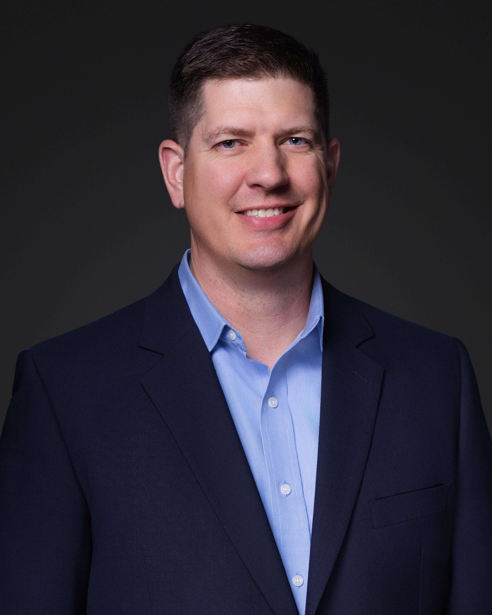 A man with short brown hair, wearing a dark suit jacket over a light blue button-up shirt, stands against a plain dark background, smiling at the camera.