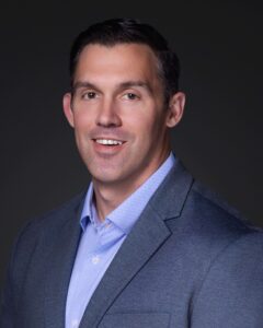 A man with short dark hair is wearing a light blue collared shirt and a gray blazer. He is smiling slightly and facing the camera against a dark, plain background.