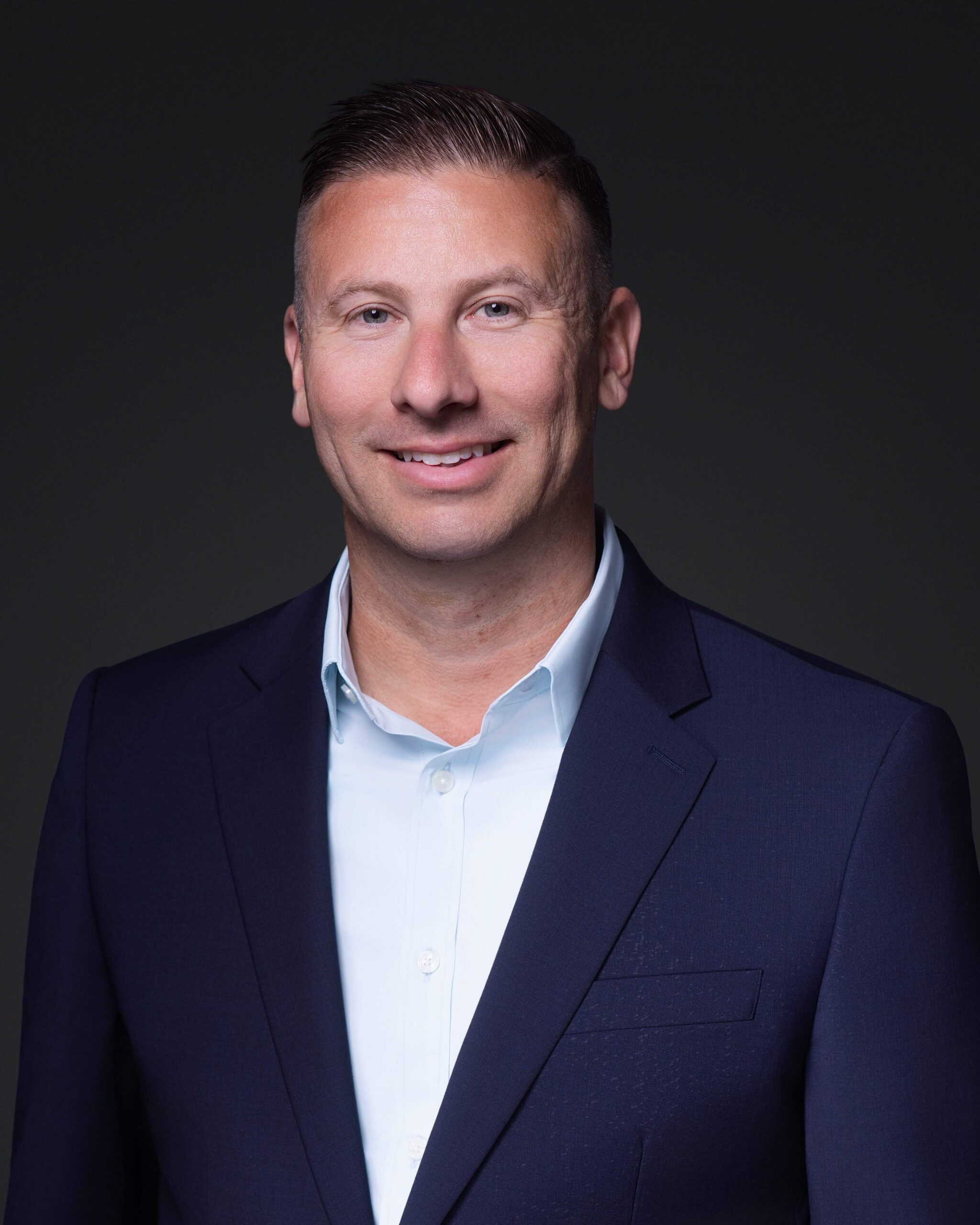 A man wearing a navy blue suit jacket and light blue dress shirt is standing against a plain, dark background. He has short hair and is smiling slightly at the camera.