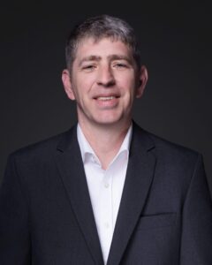 A man with short gray hair is wearing a dark suit jacket and a white collared shirt, smiling slightly in front of a dark background.