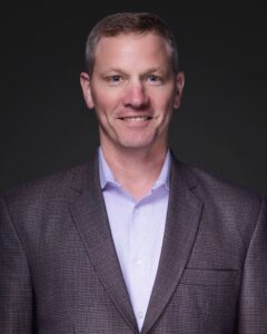 A man with short light brown hair wears a light purple shirt and a checked blazer, posing and smiling slightly in front of a dark, plain background.