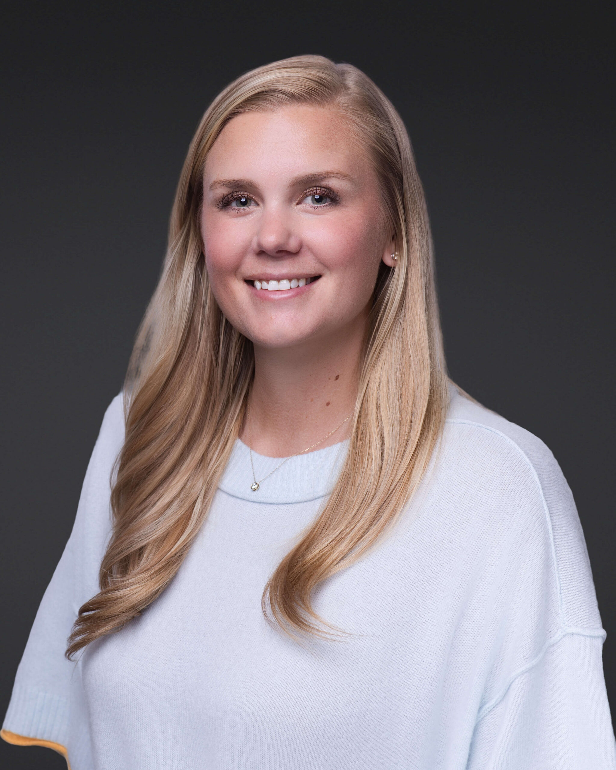 A woman with long blonde hair wearing a light-colored sweater and a small necklace smiles at the camera against a dark gray background.